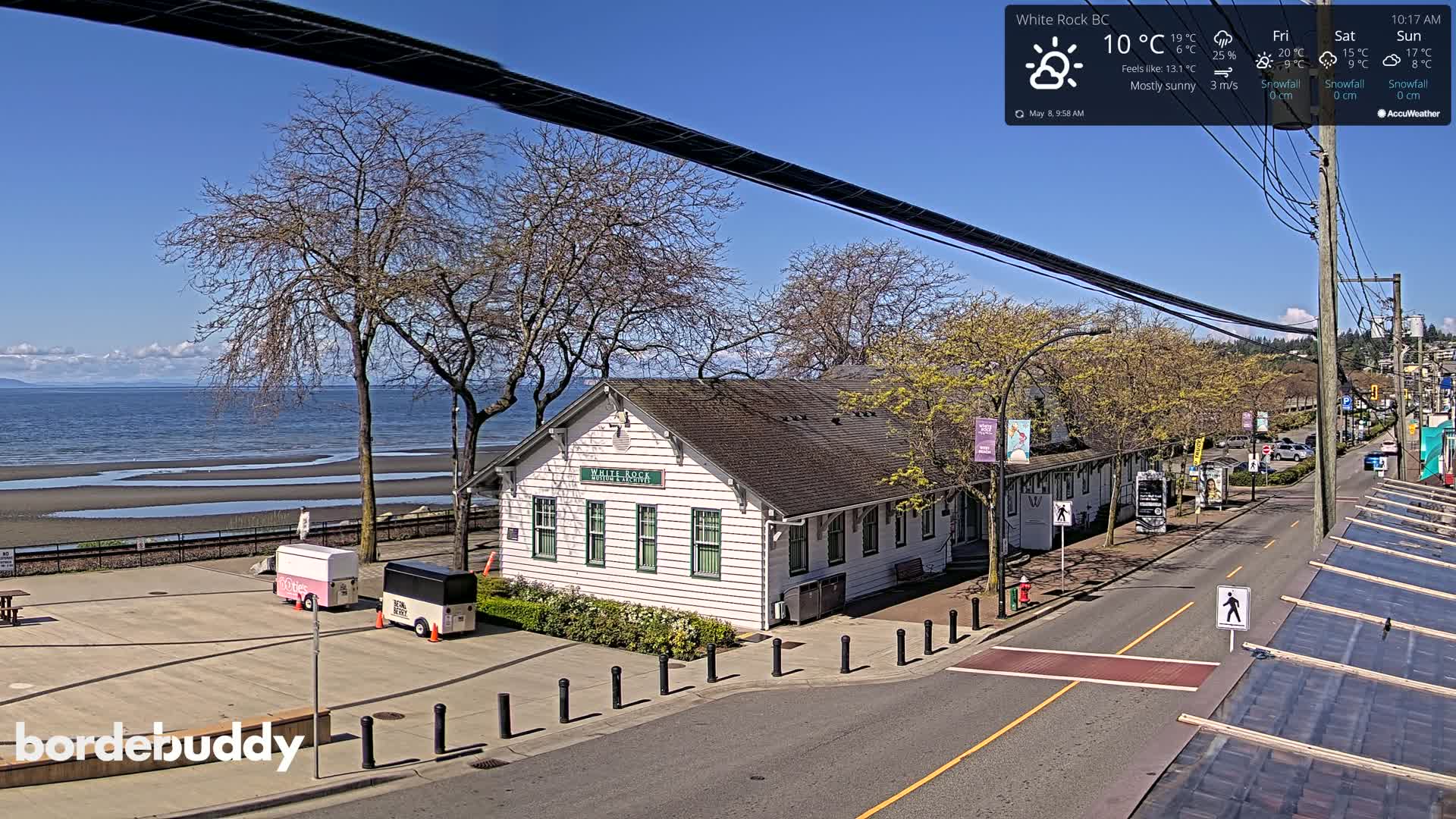 A mostly sunny day reveals a white building near a beach with a calm ocean, trees, and a street with parked cars.