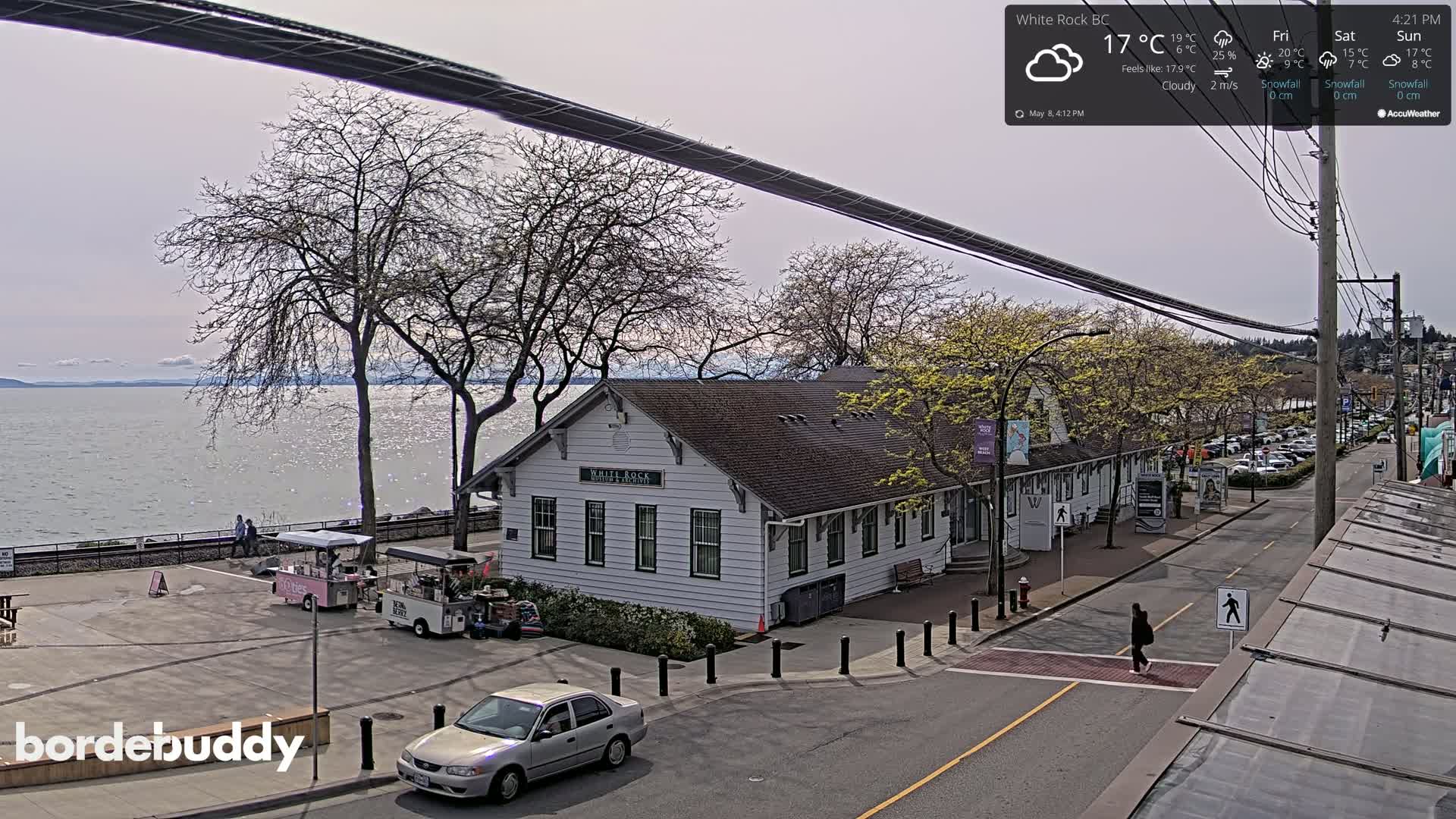 A mostly cloudy, sunny day overlooks a waterfront town with a white building, parked cars, and a person crossing the street.