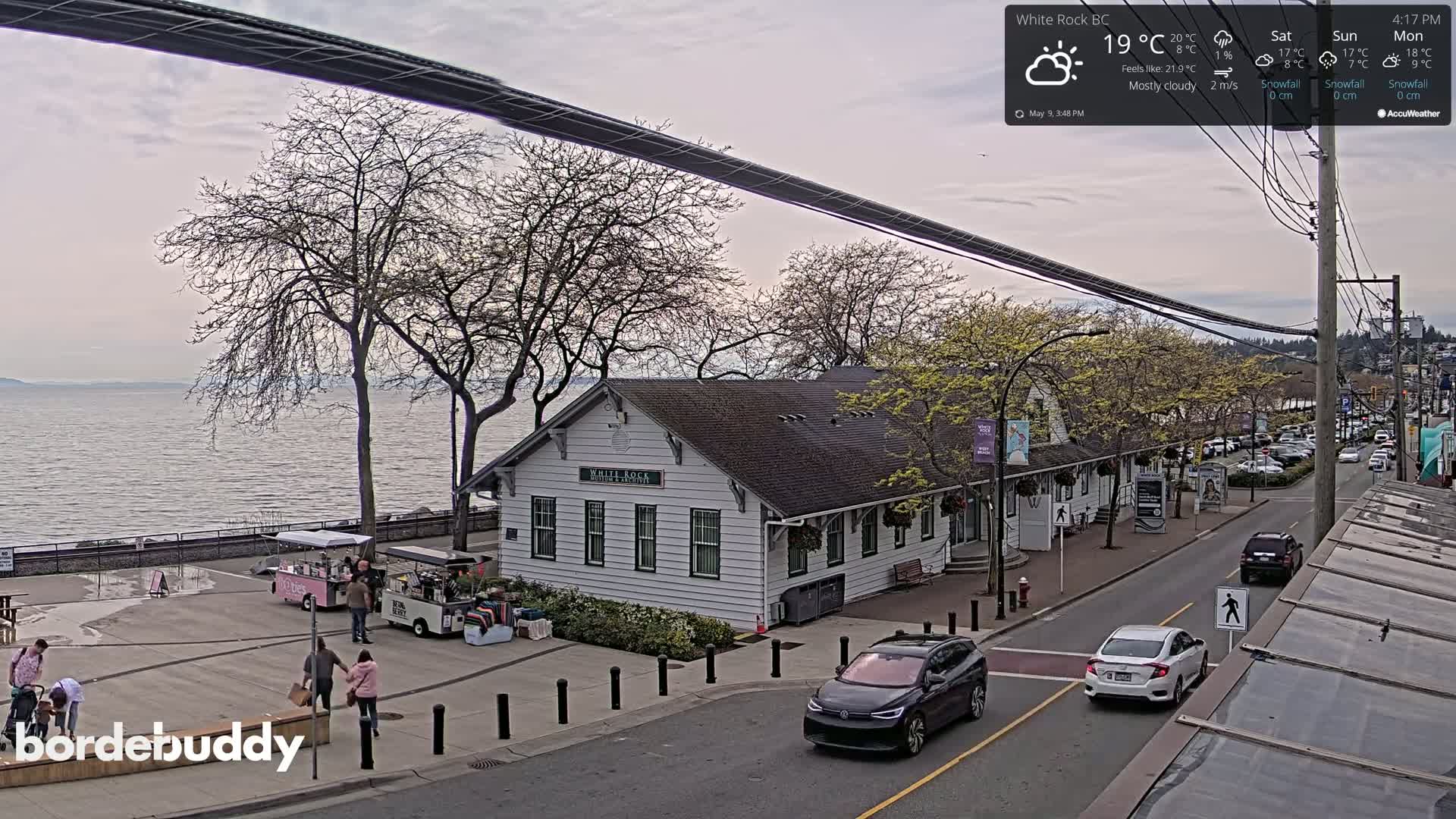 A mostly cloudy day reveals a waterfront scene with a white building, cars on a street, and people near food trucks, all overlooking a calm body of water.