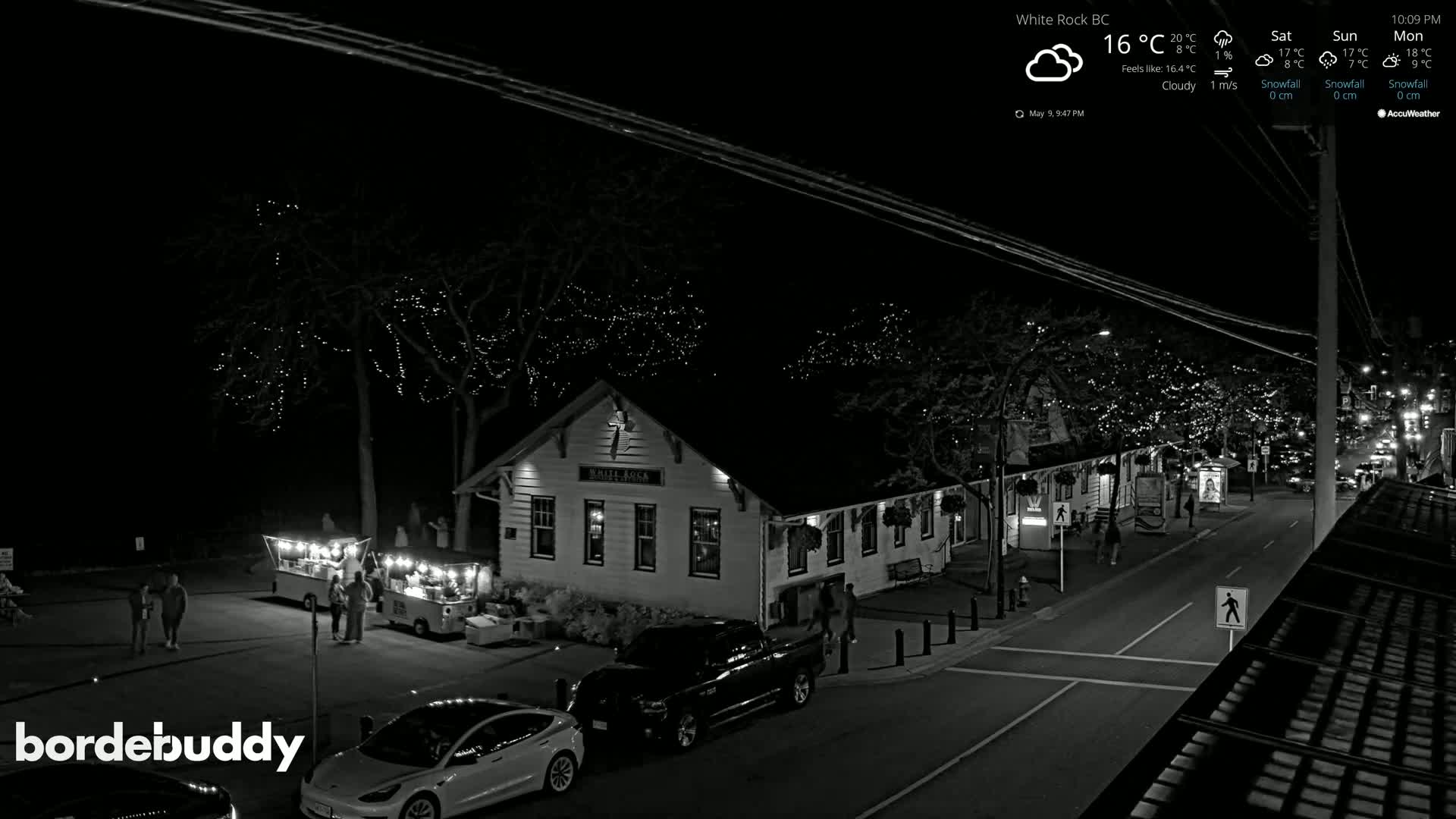 A nighttime, cloudy scene shows a street with a building illuminated by string lights, several parked cars and a few pedestrians.