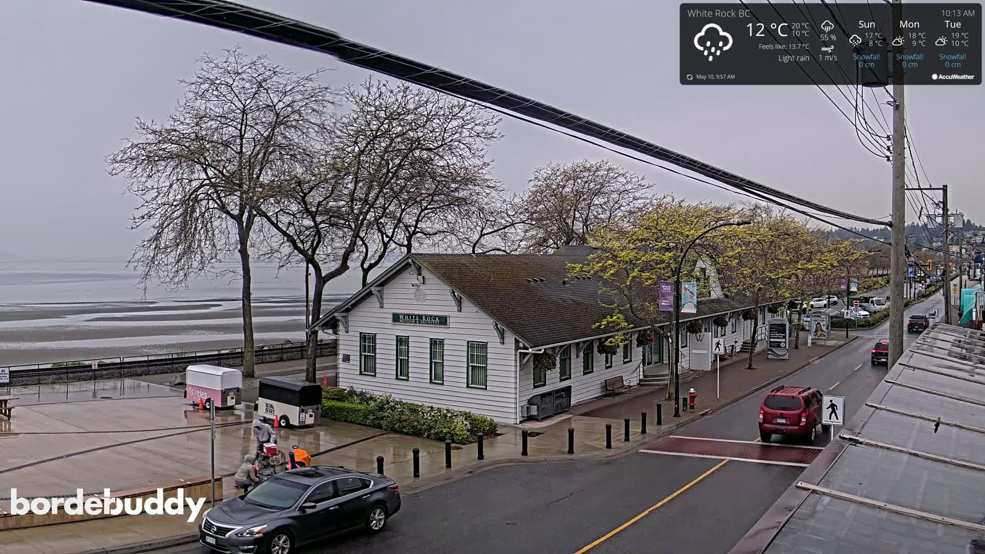 A wet, overcast street scene shows a white building near a shoreline with a few people and cars; trees are bare with some budding leaves.
