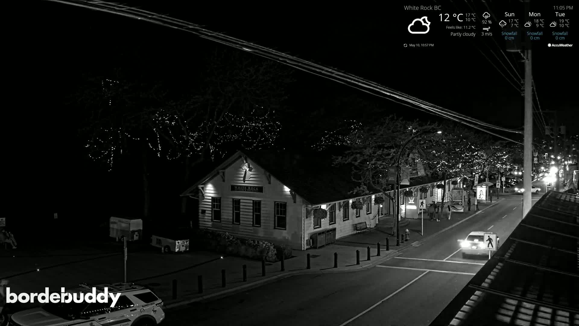 A nighttime, partly cloudy scene shows a street with a building illuminated, streetlights, cars, and pedestrians, with string lights visible in the trees.