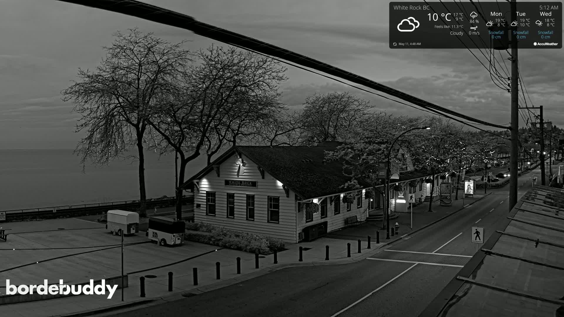 A grayscale, overhead view shows a seaside town at dusk under cloudy skies, featuring a building near the water's edge and a street lined with trees.
