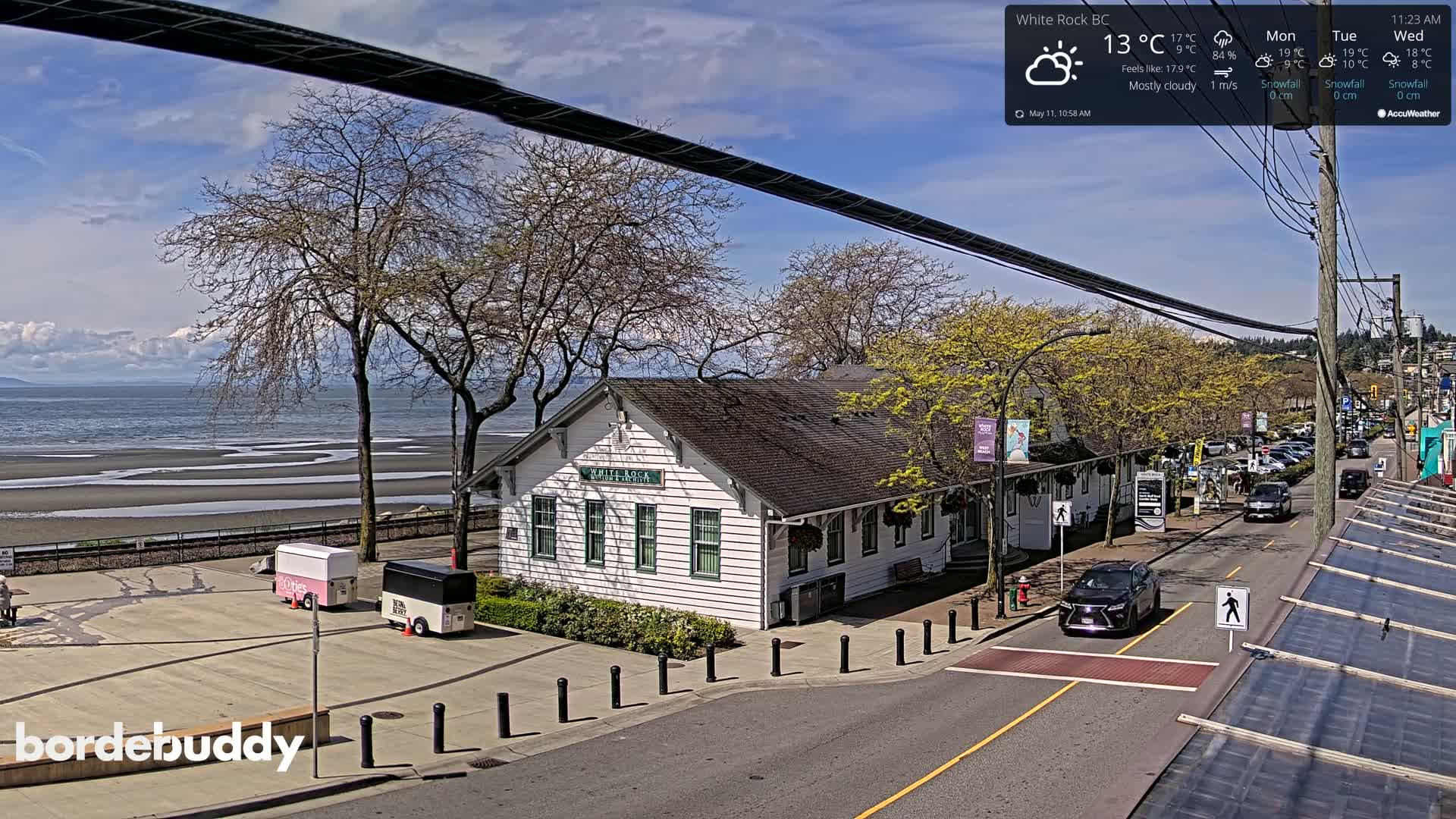 A mostly cloudy, 13°C day overlooks a white building near a beach and a street with cars parked along the side.
