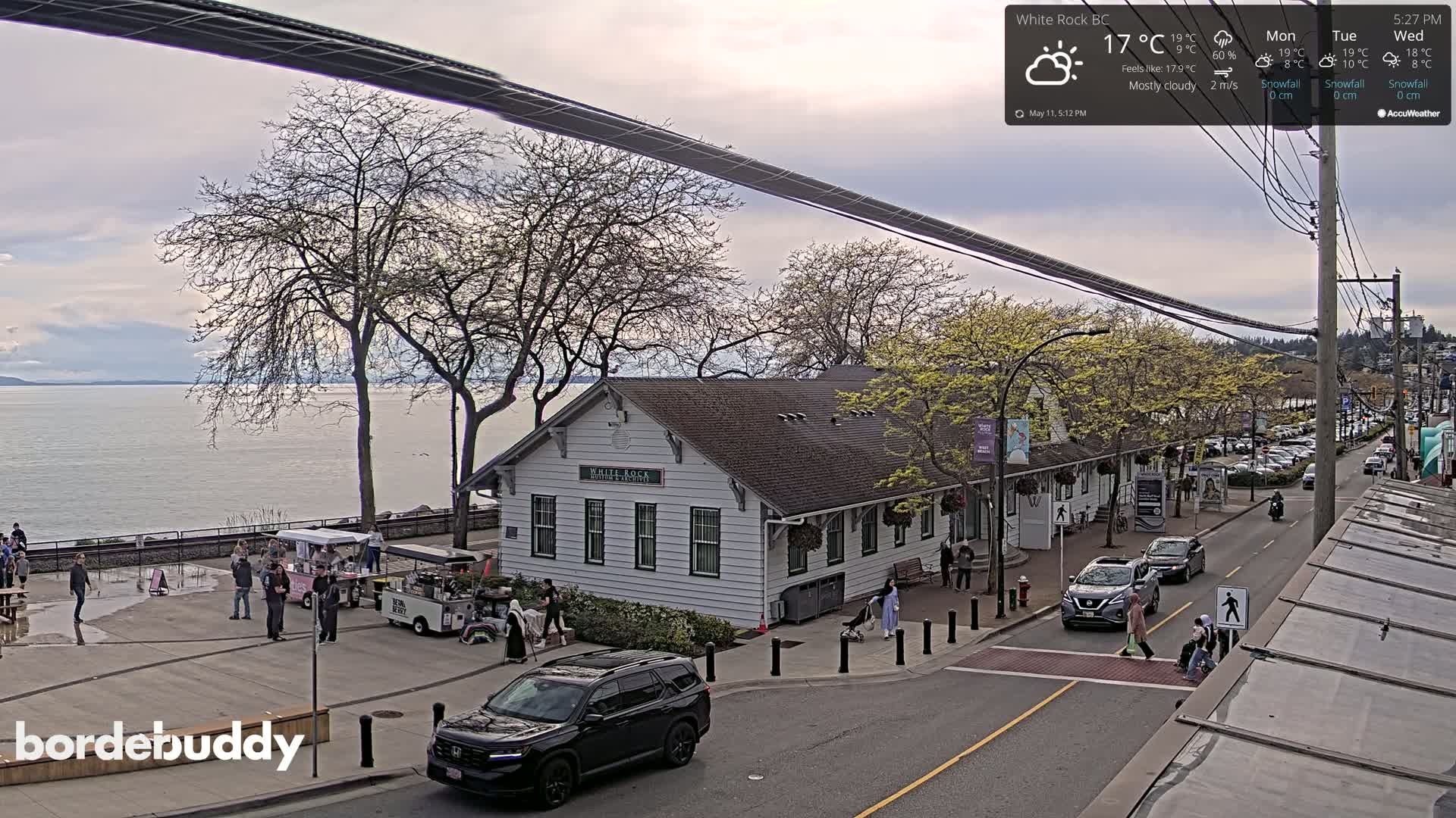 A mostly cloudy, cool day overlooks a waterfront town with a white building, several people, cars, and food stands near a calm body of water.