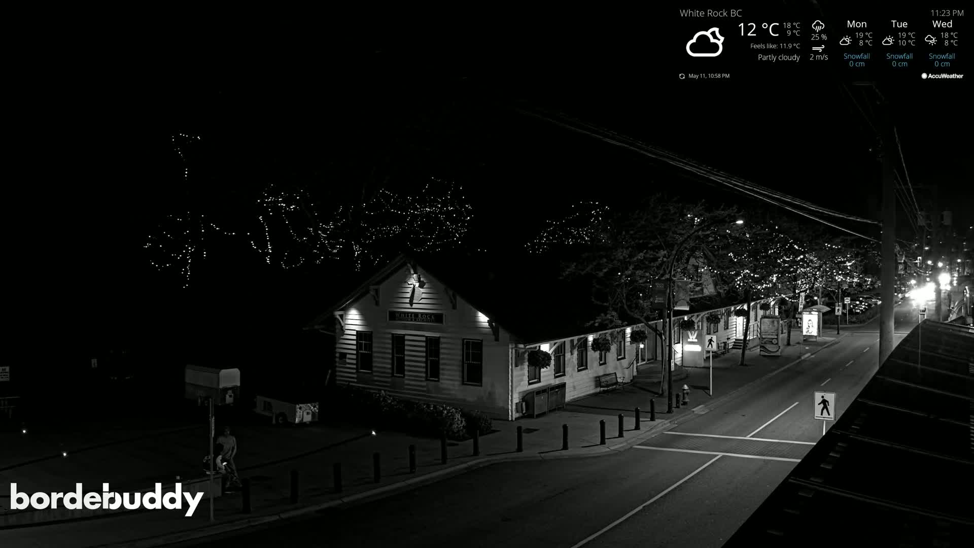 A partly cloudy night scene shows a street with a building illuminated, trees strung with lights, and a person walking.