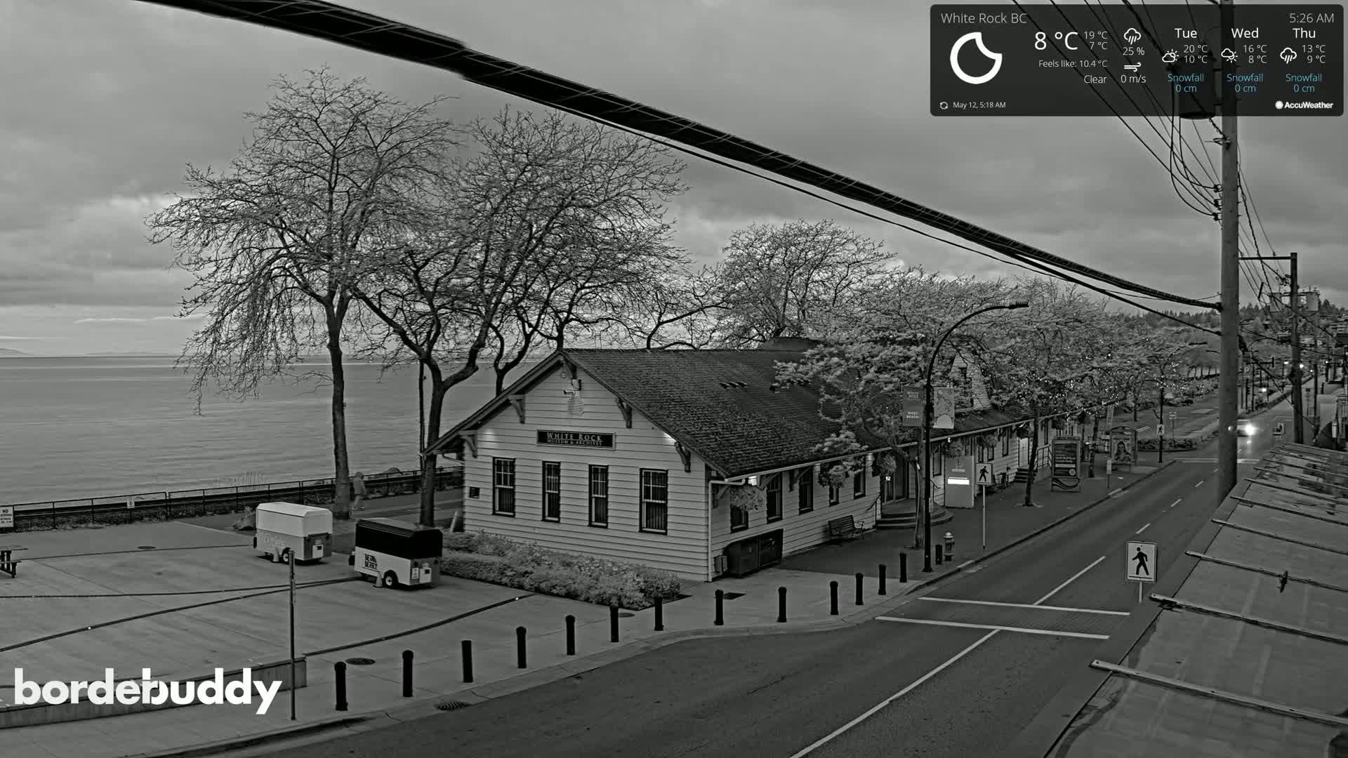 A grayscale image shows a waterfront street scene with a building, bare trees, parked vehicles, and calm water under an overcast sky.