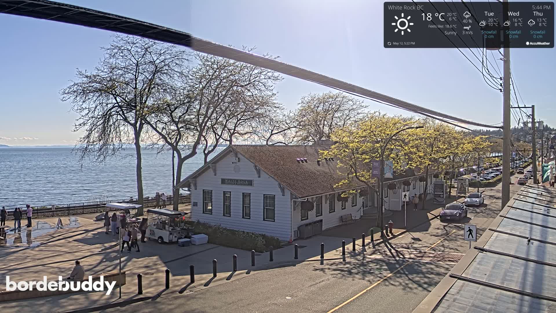 A sunny waterfront scene shows a white building near a calm body of water, with trees, people, and parked cars lining a street.