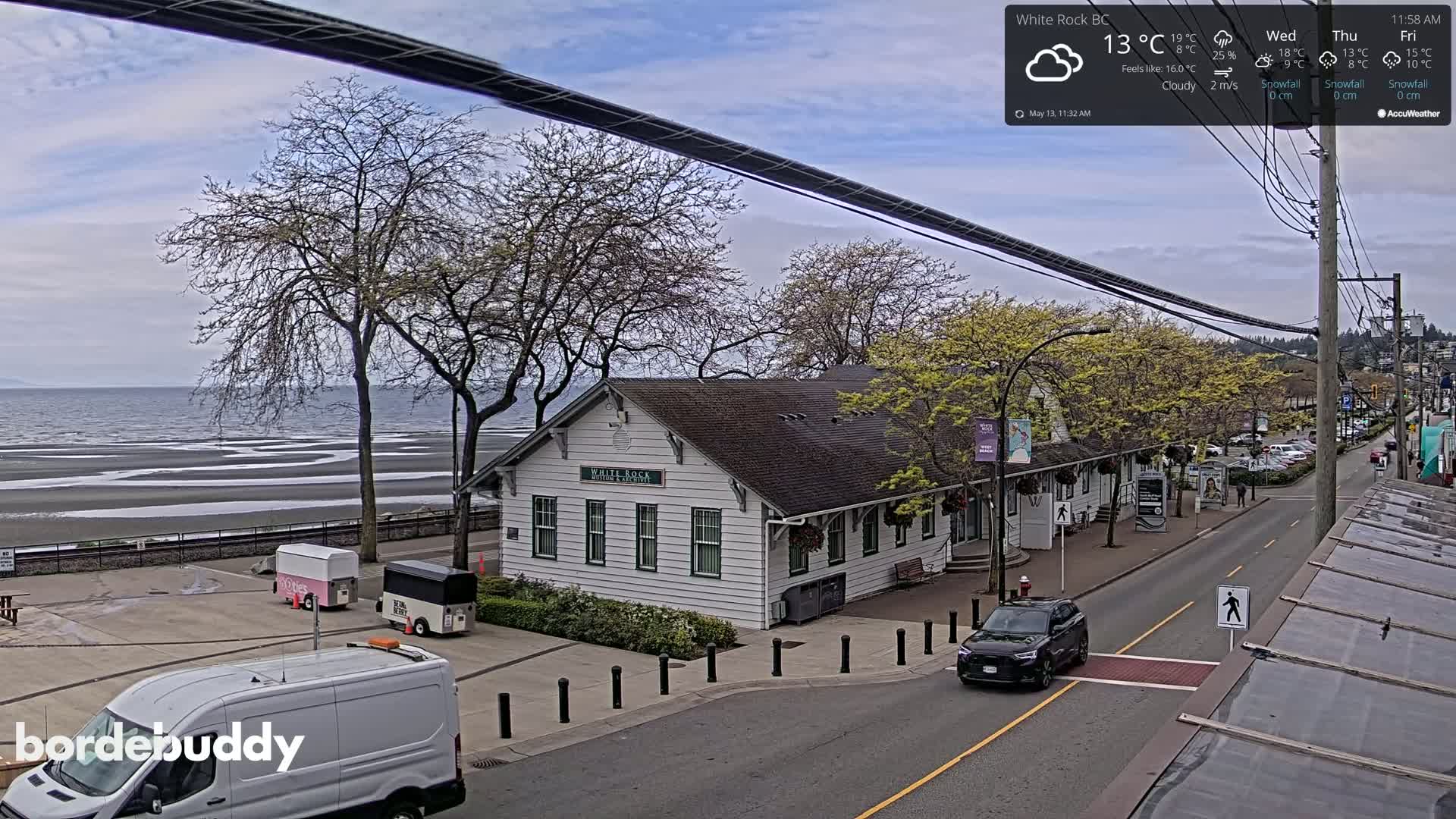 A cloudy day overlooks a waterfront street scene with a white building, parked cars, and food trucks near a calm ocean.