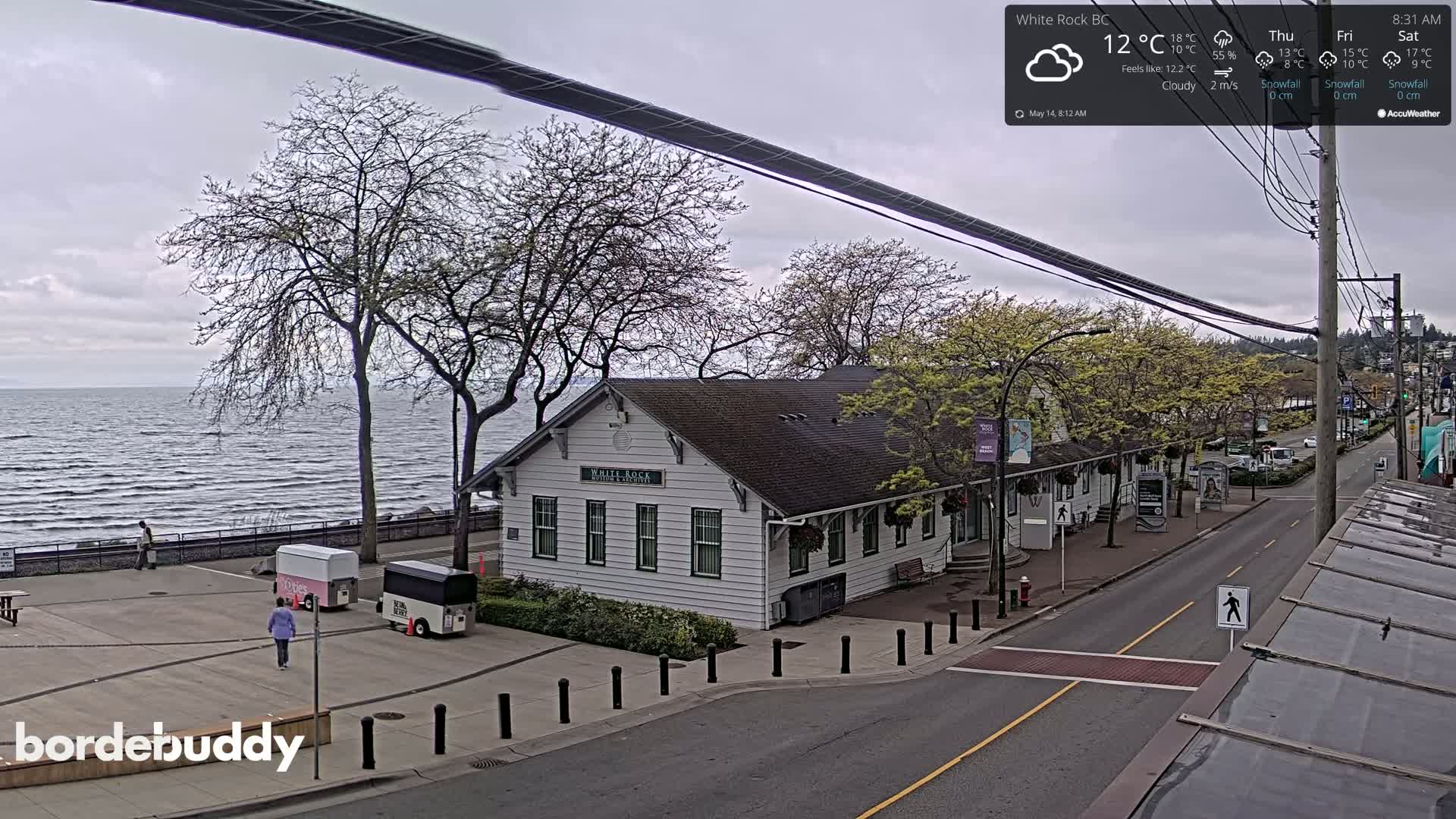 A cloudy day overlooks a waterfront street scene with a white building, bare trees, and a calm body of water.