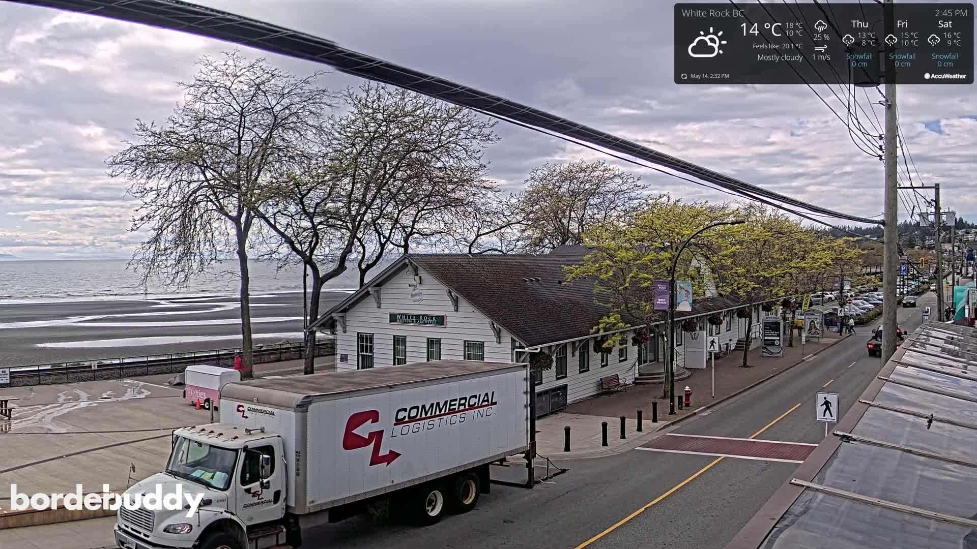 A mostly cloudy day reveals a seaside town with a white building, a parked delivery truck, and a calm ocean visible in the background.