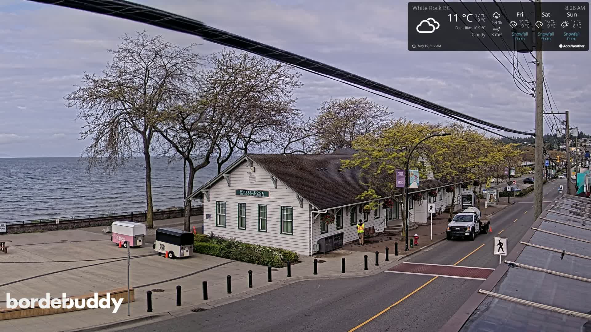 A cloudy day shows a waterfront view of a white building near a road with parked vehicles and food carts, overlooking a calm body of water.