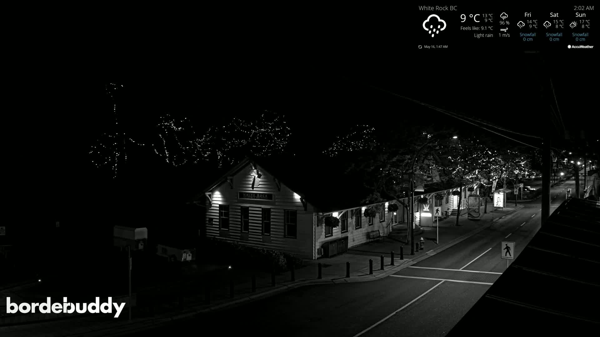 A nighttime, lightly raining scene shows a street with a building illuminated, string lights in trees, and a road.