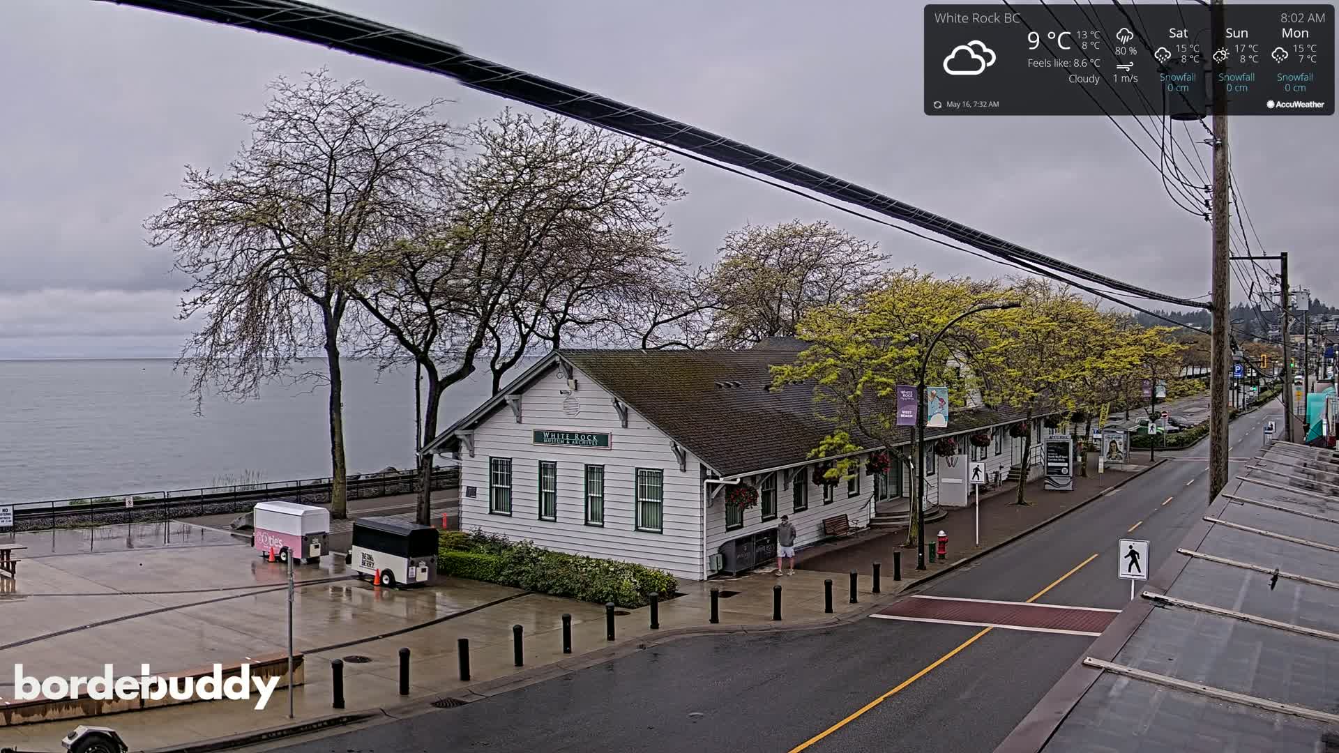 A cloudy day shows a waterfront view of a white building near a calm body of water with a street and some trees.