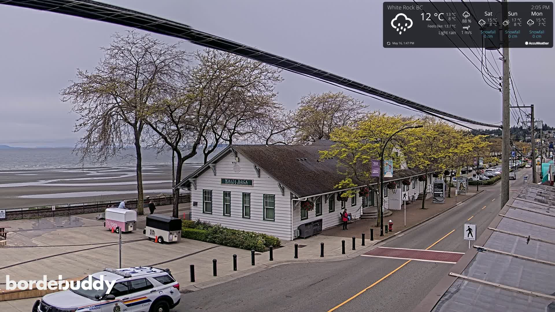 A light rain falls on a seaside town with a white building, trees budding with green leaves, a beach, and a police car parked on the street.