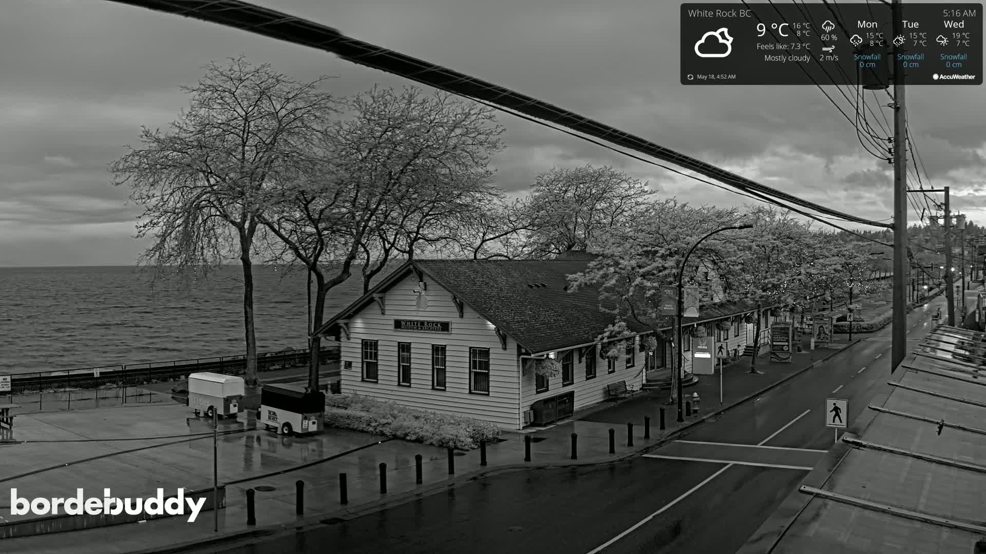 A mostly cloudy, wet day reveals a seaside town with a white building, bare trees, and a calm body of water.