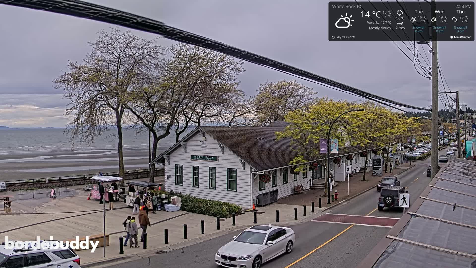 A mostly cloudy day reveals a seaside town with a white building near a beach, cars on a street, and people walking near food vendors.