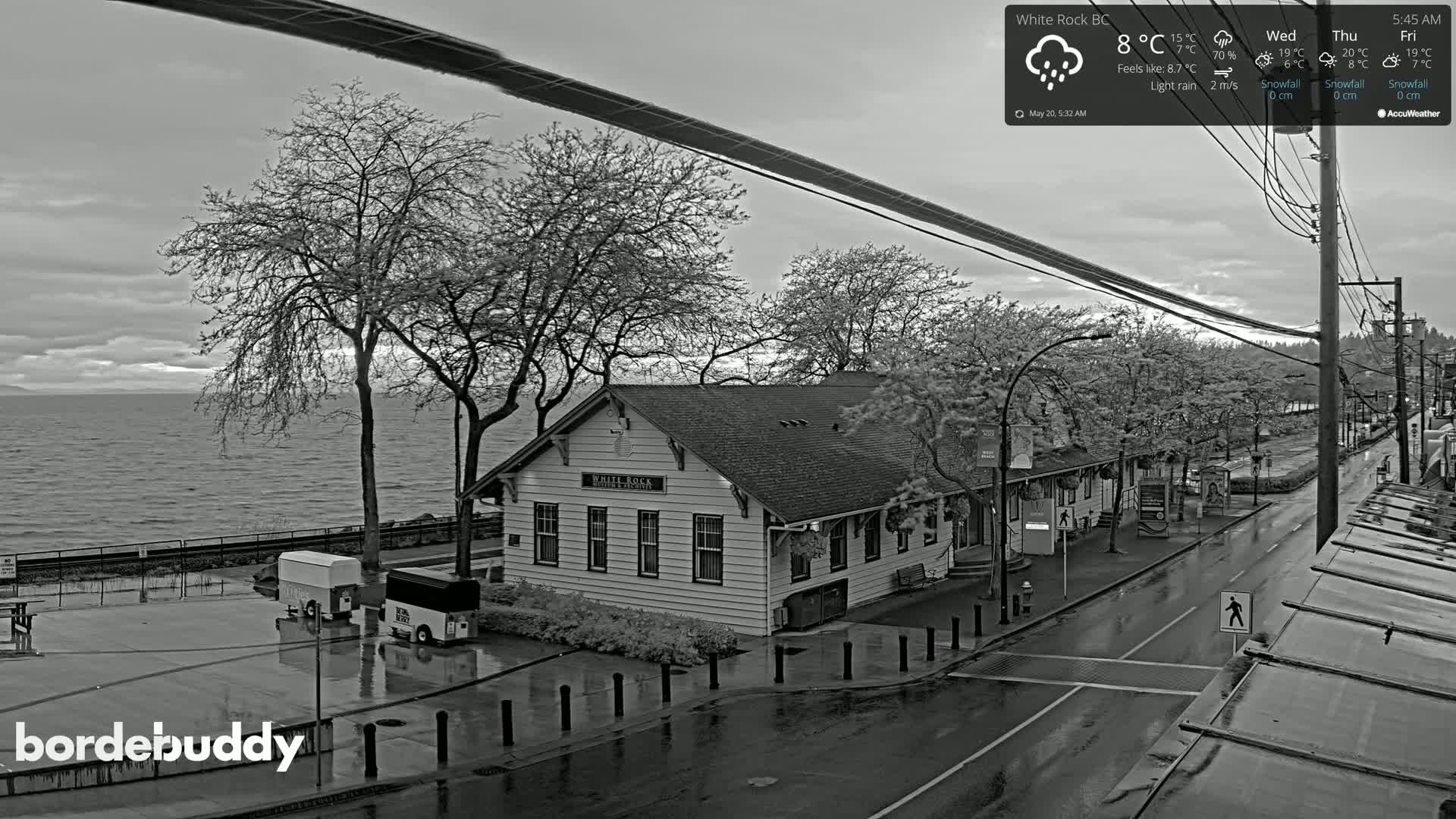 A black and white, high-angle view of a seaside town on a rainy day shows a long, low building near the water, with wet streets and parked vehicles.