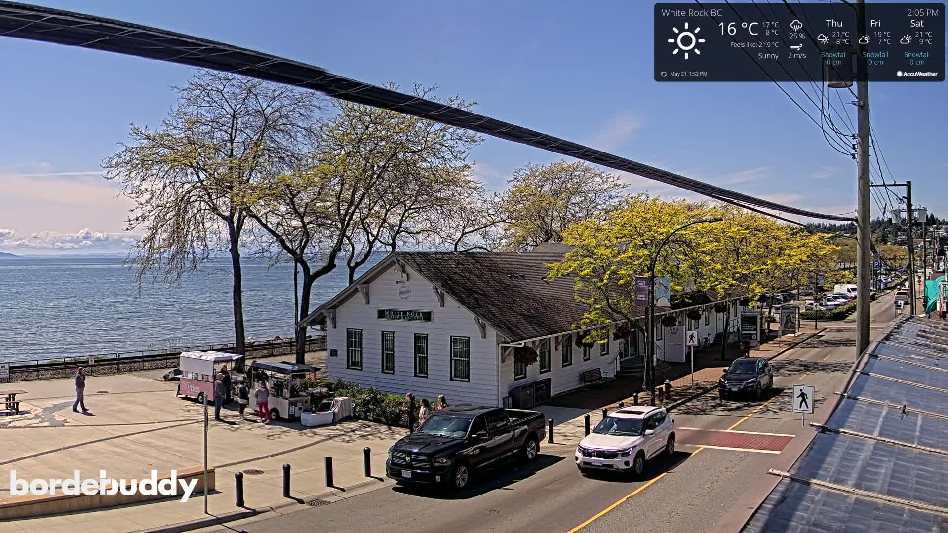 A sunny waterfront scene shows a white building near a street with parked cars and food trucks, with trees in bloom and a calm body of water beyond.