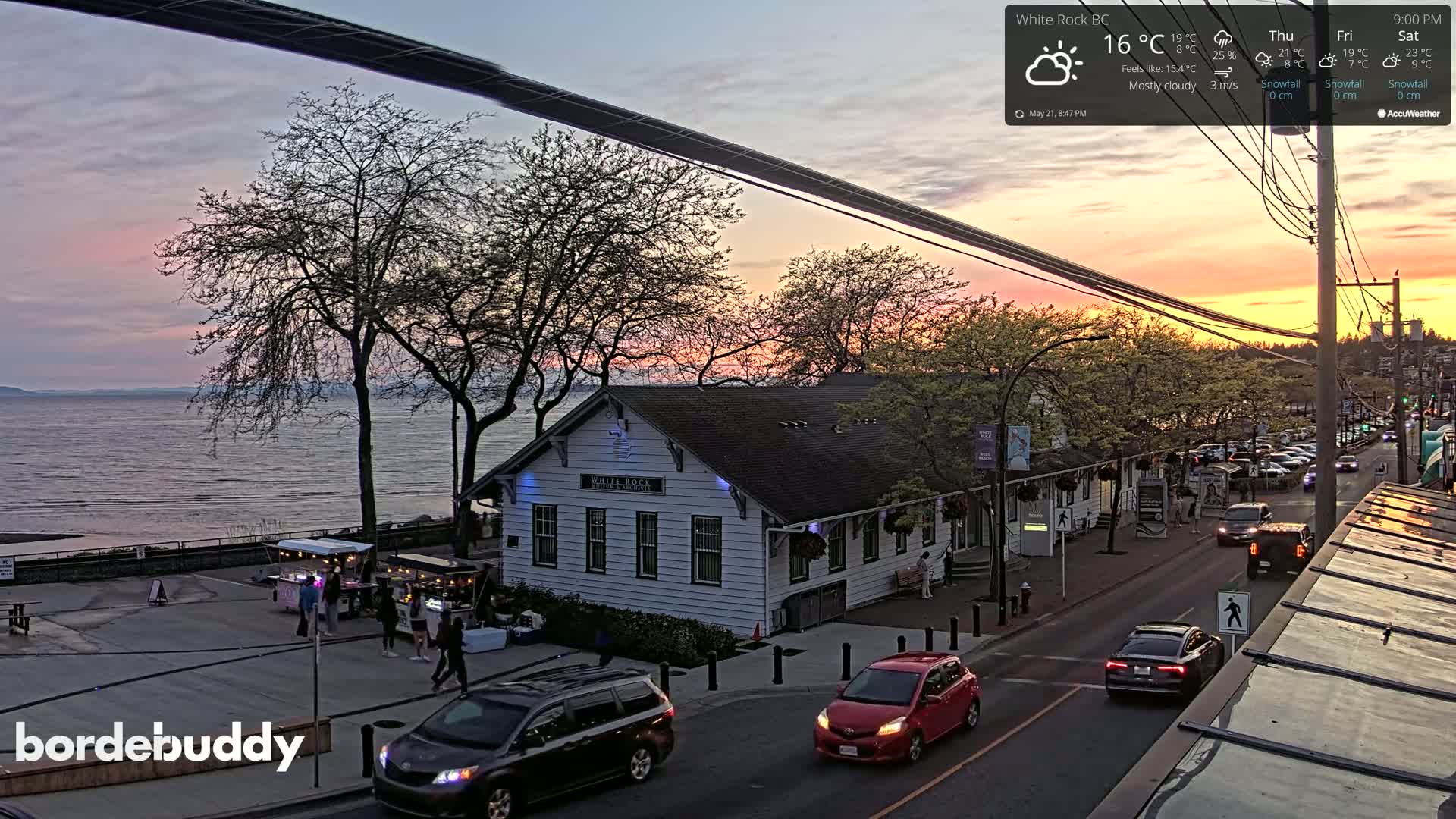 A mostly cloudy sunset over a calm body of water, viewed from above a street with cars and pedestrians near a white building and food vendors.