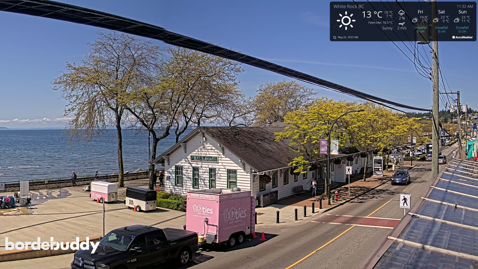 A sunny waterfront scene shows a white building near a roadway with several vehicles and pedestrians, along with food trucks and trees budding with new leaves.