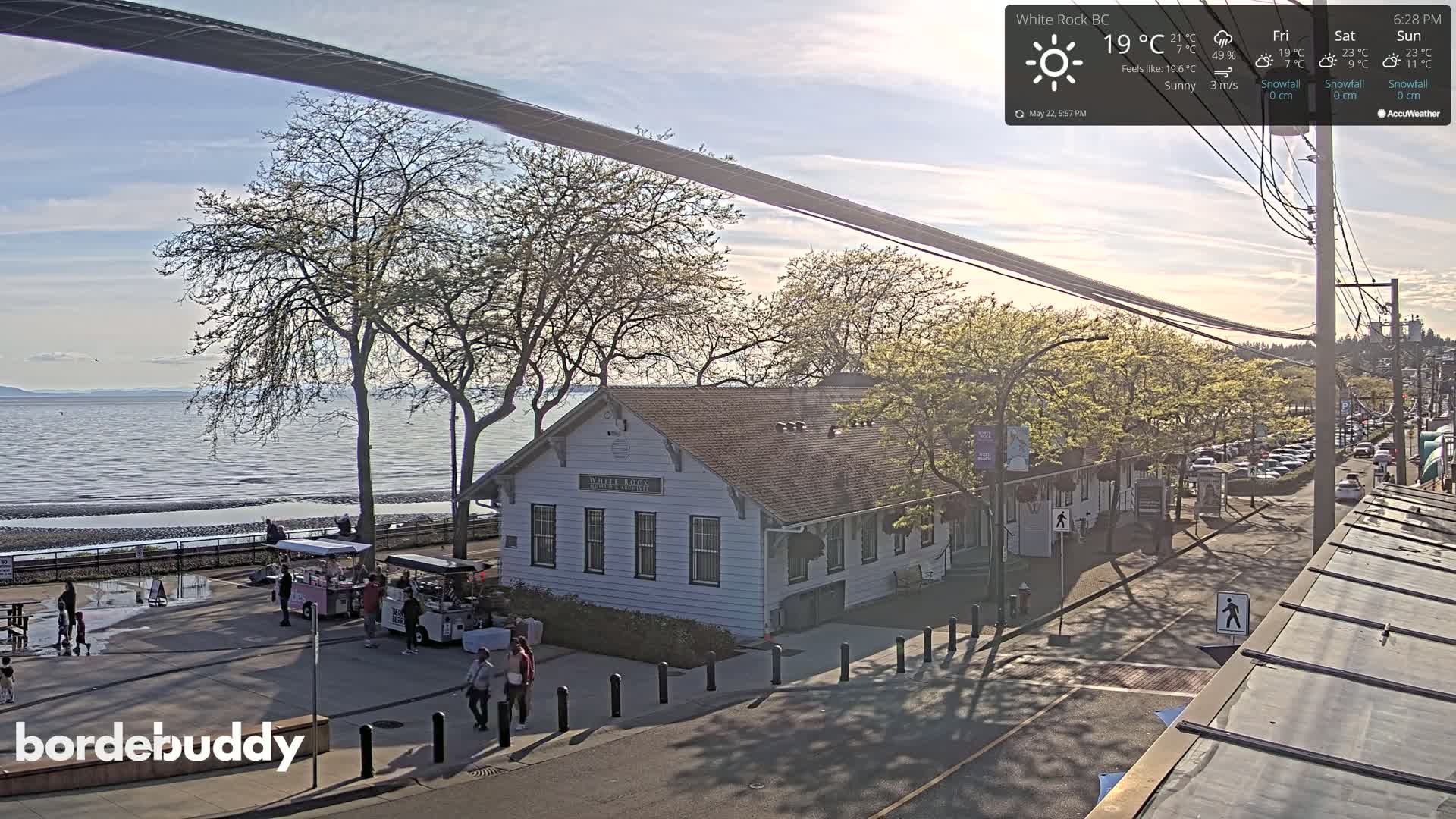 A sunny waterfront scene shows a white building near a street with parked cars and people walking, trees, and a calm body of water beyond a paved shoreline.