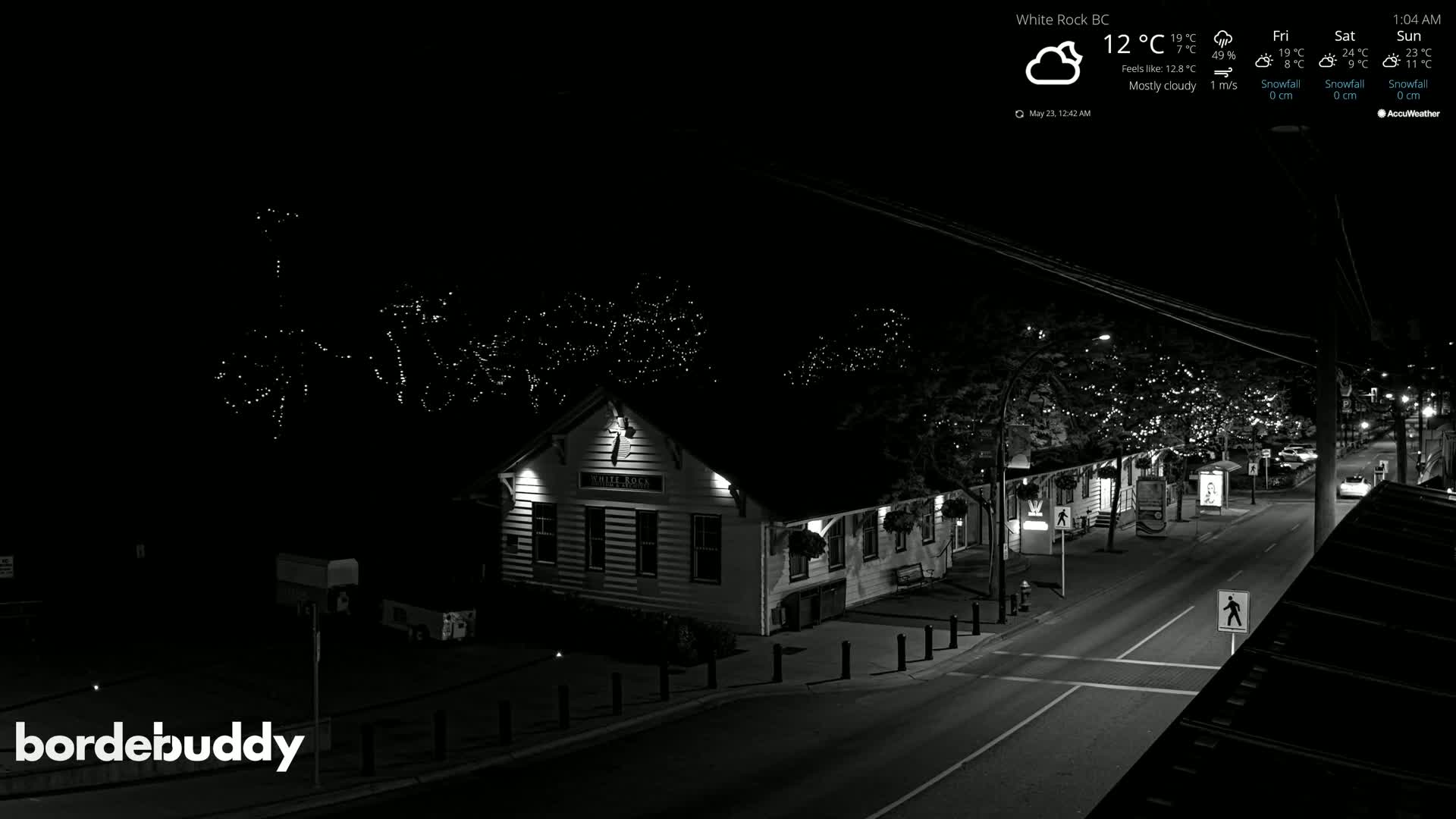 A mostly cloudy night scene shows a street with a building decorated with lights, and a few cars parked on the road.