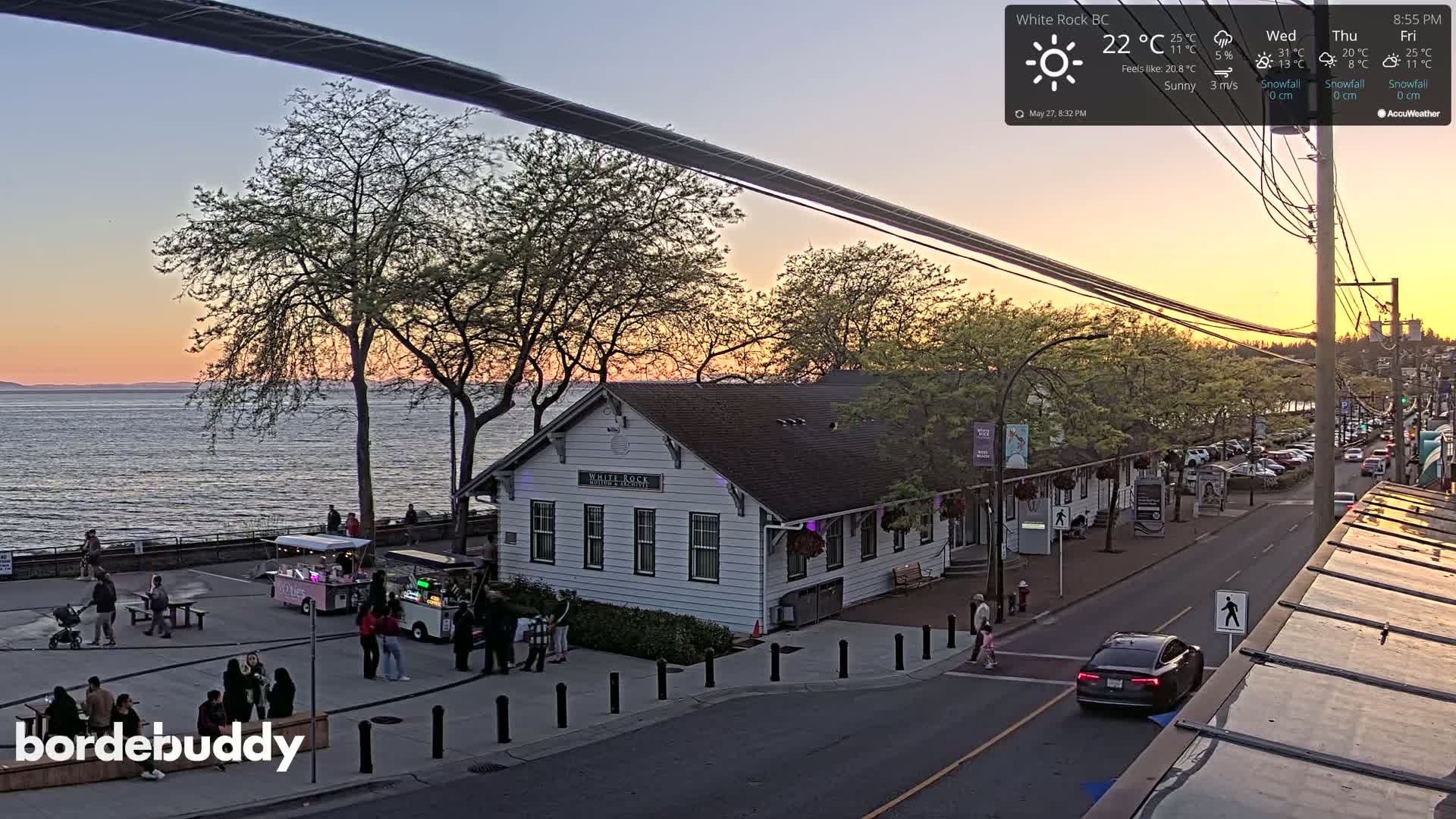 A waterfront street scene at sunset shows a white building, food trucks, people gathered in a plaza, parked cars, and a calm body of water in the background.