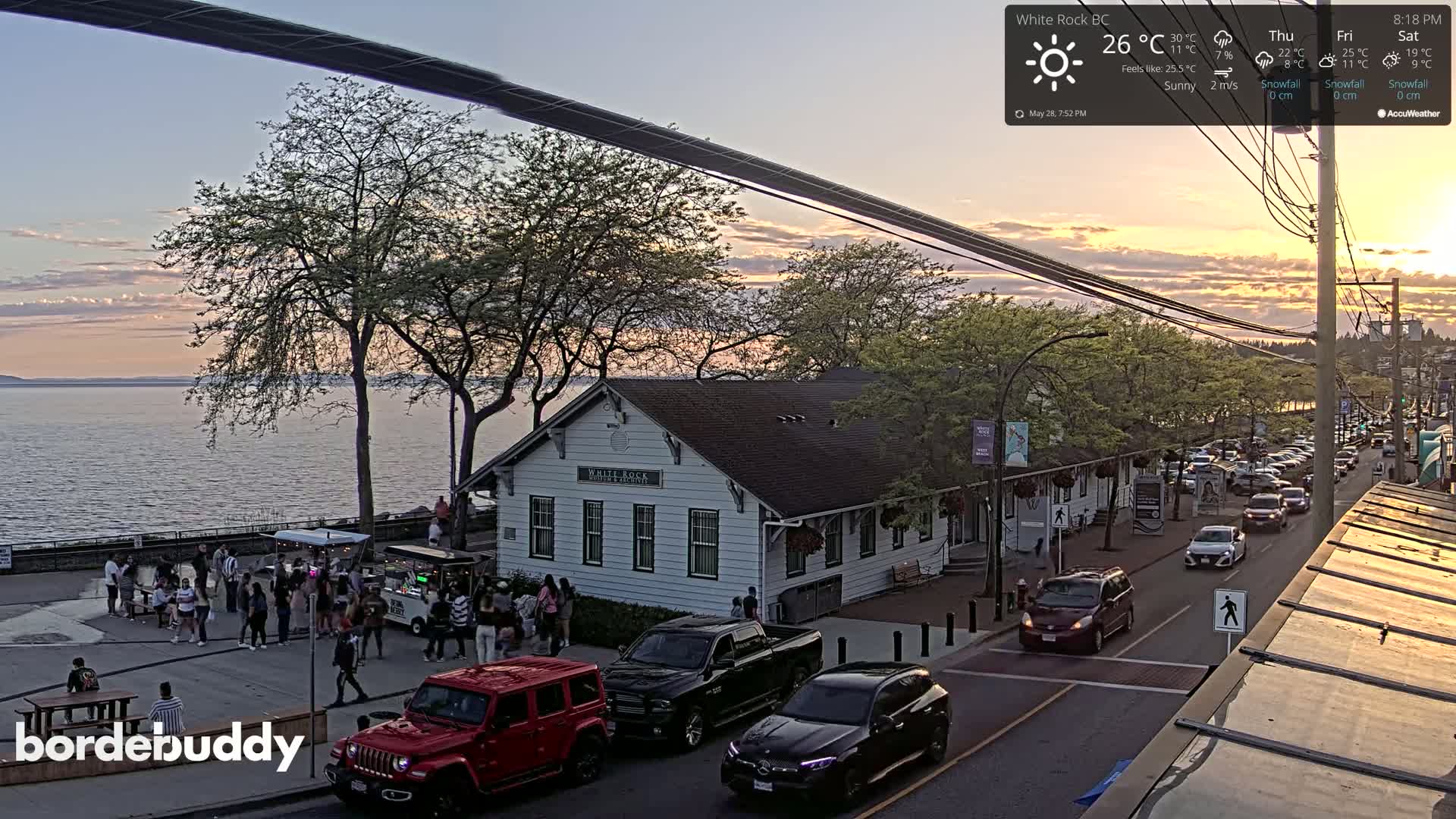 A waterfront town at sunset, with people gathered near a building and cars parked along a street.