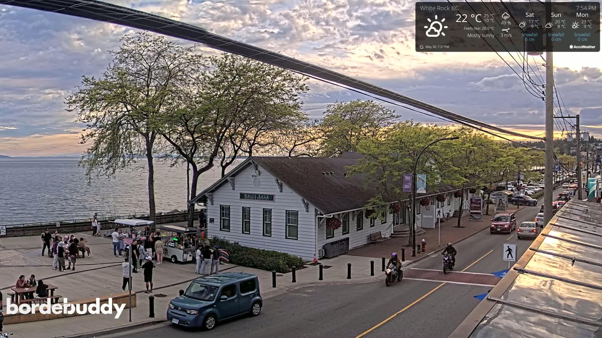 A mostly sunny sunset over a calm body of water, viewed from above a waterfront town with a white building, people gathered in a plaza, and cars driving on a street.