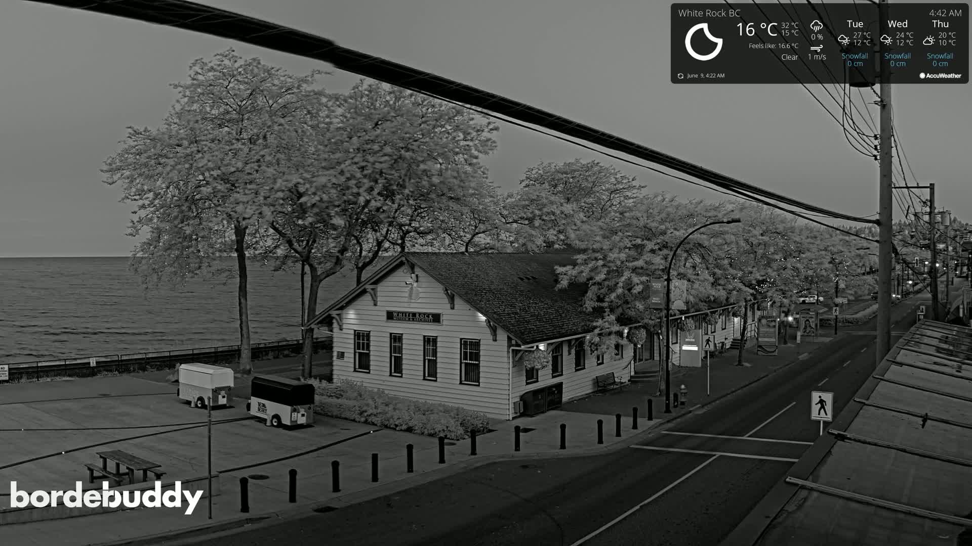 A grayscale image shows a waterfront view of a building, trees, a street, and calm water under a clear sky.