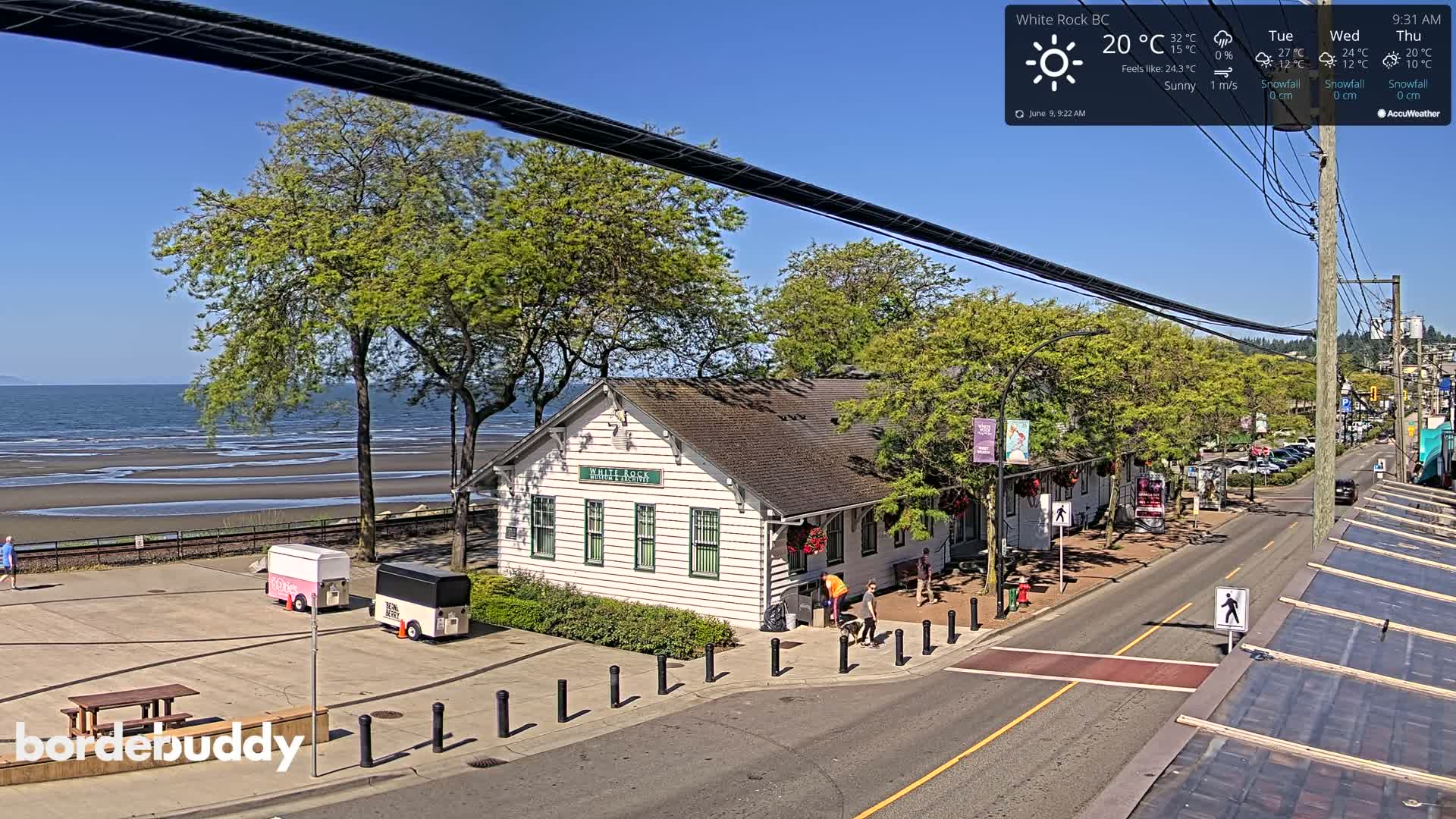 A sunny seaside town scene shows a white building near a beach with calm water under a clear blue sky.