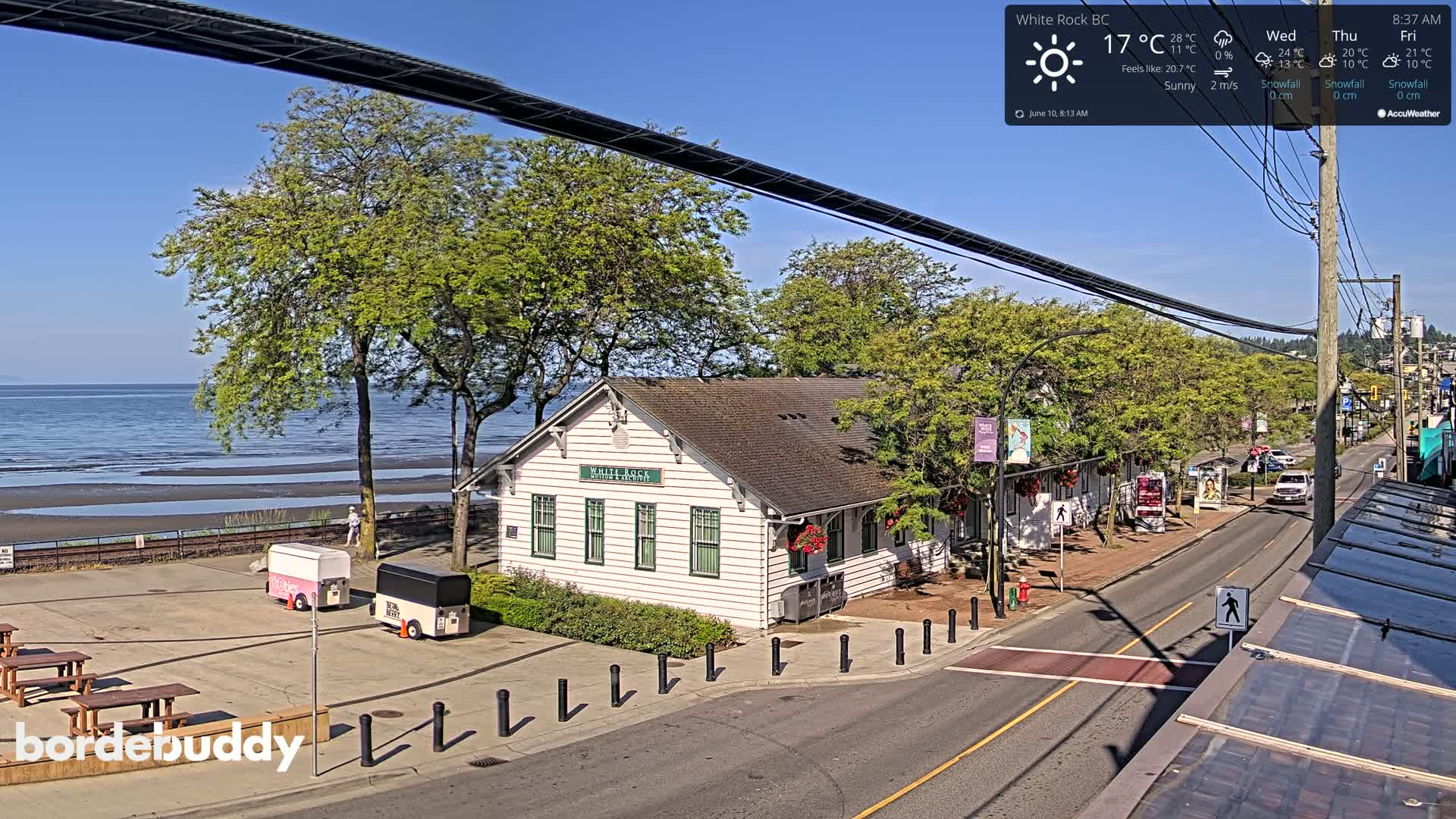 A white building sits near a calm ocean under a sunny sky, next to a street with cars and trees.