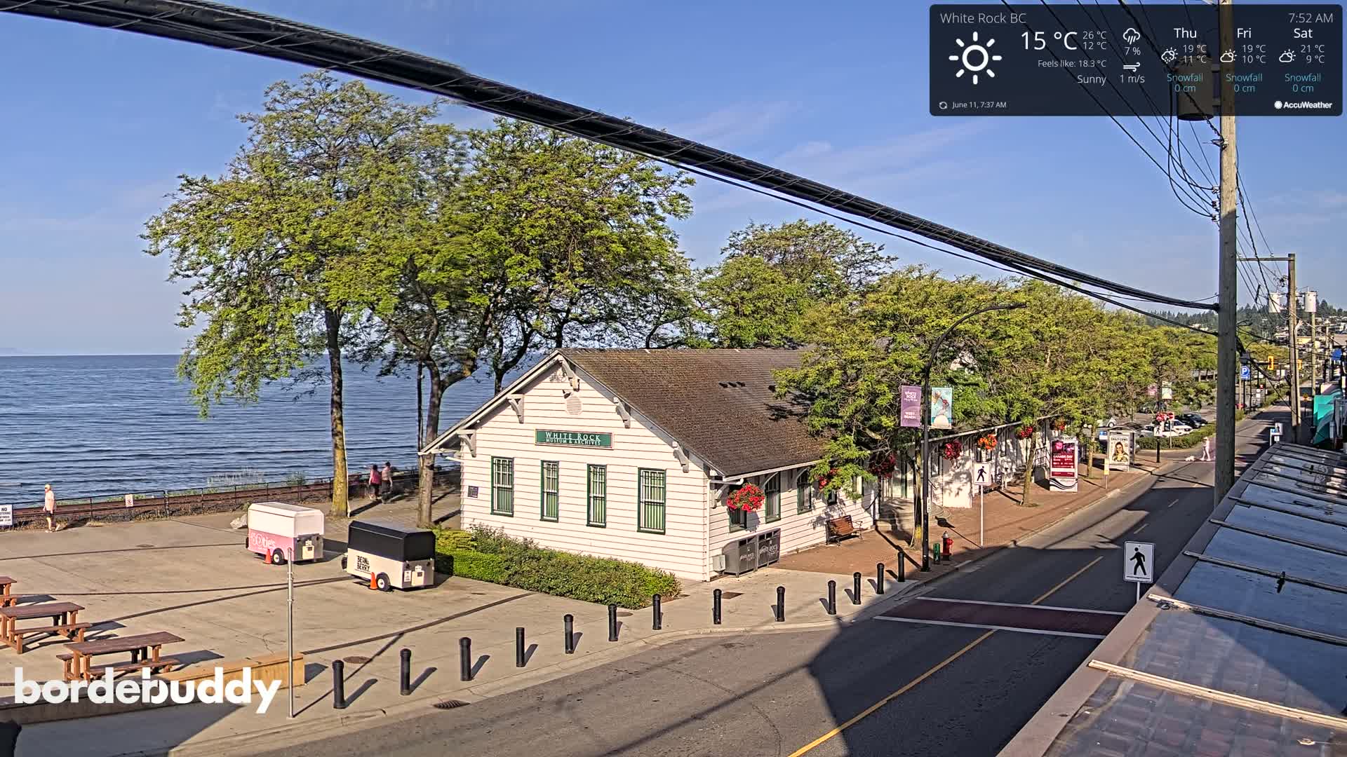 A white building sits near a calm ocean under sunny skies, with food carts and a few pedestrians visible in a town square.