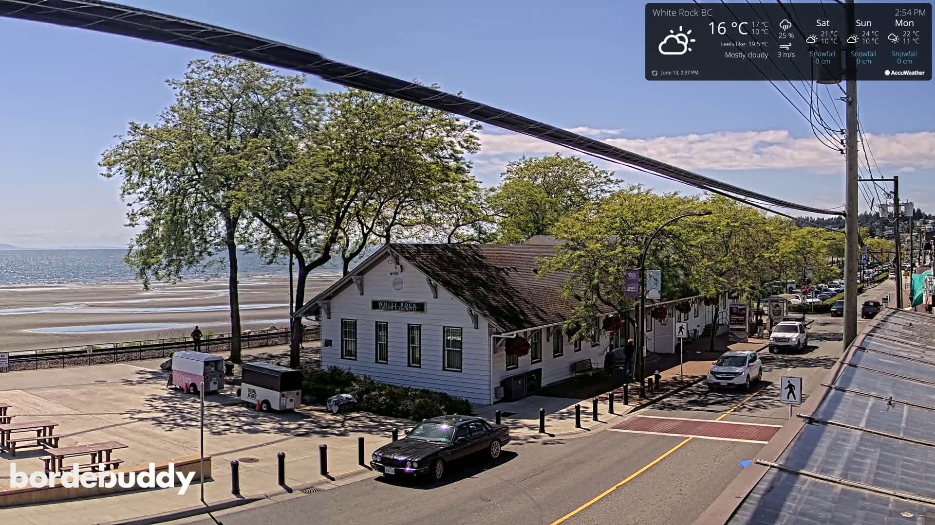 A mostly cloudy day shows a seaside town with a white building near a beach, cars on a street, and trees.