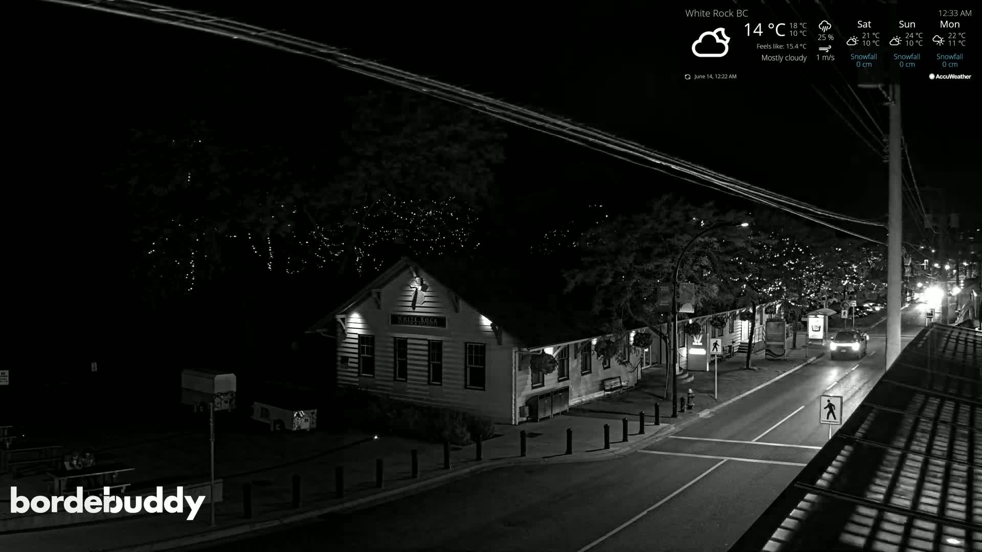 A mostly cloudy night scene shows a street with a building and some cars, with string lights visible on trees in the background.