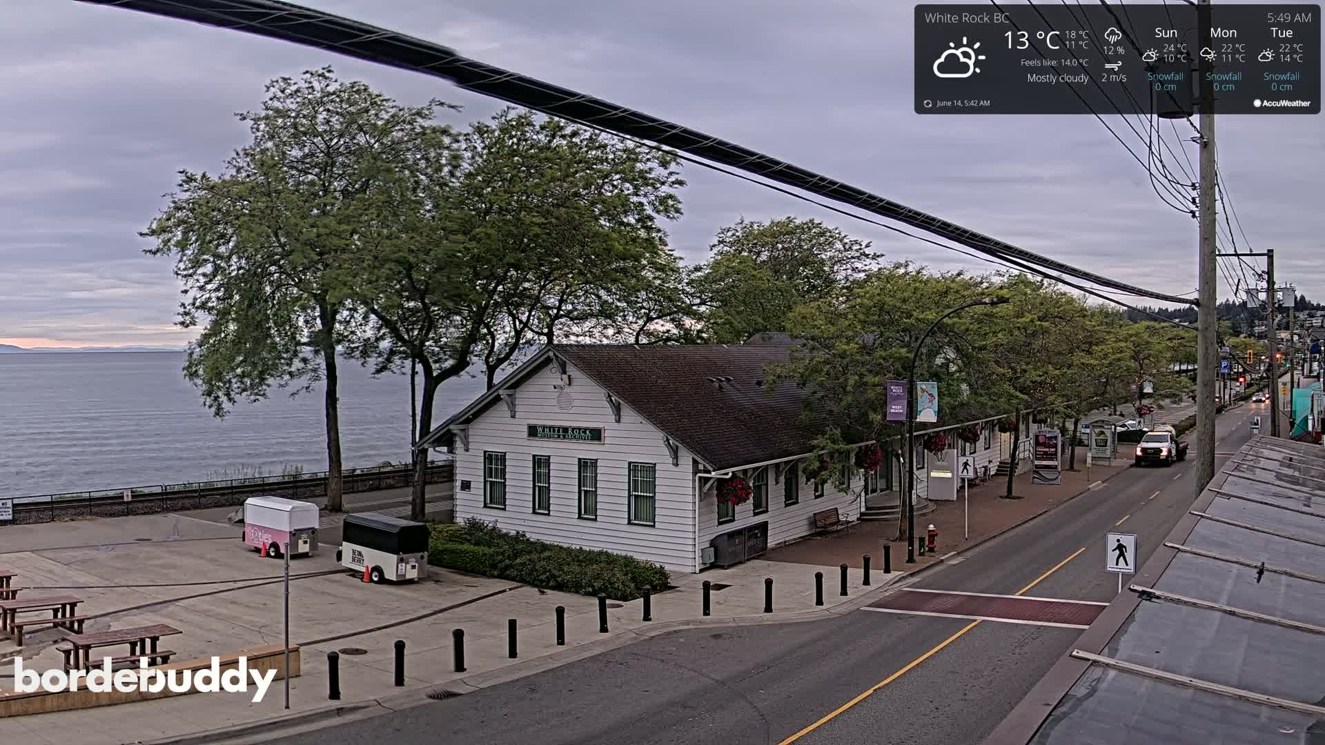 A mostly cloudy view of a waterfront town with a white building, food carts, and a street shows a calm body of water in the background.