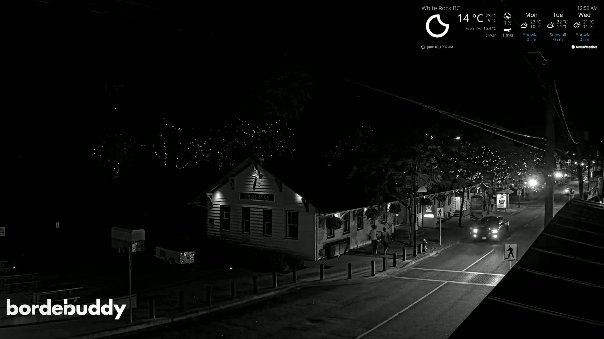 A nighttime, clear-weather view of a street scene featuring a building with lights, trees with string lights, and a few pedestrians and vehicles.