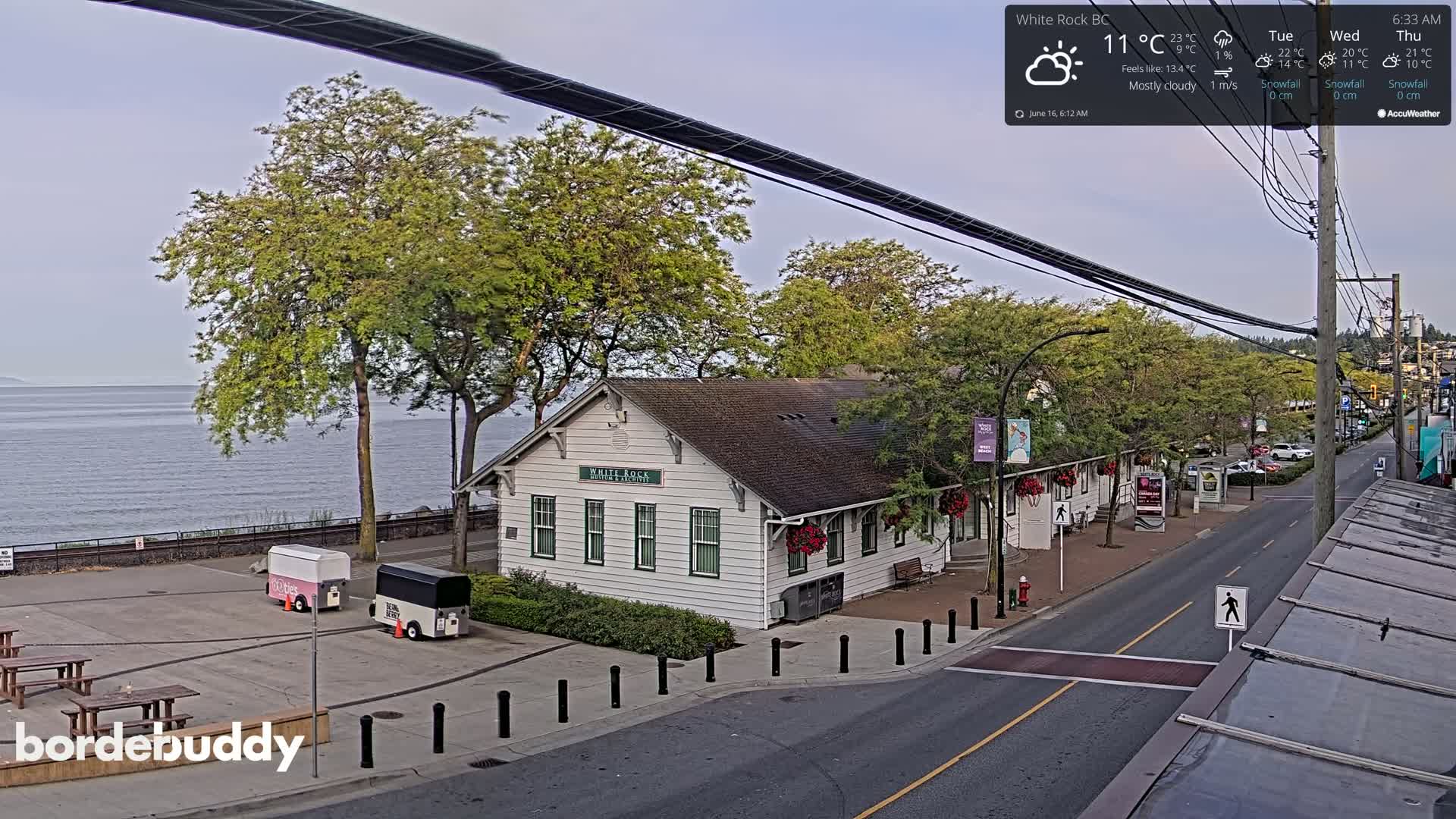 A mostly cloudy morning overlooks a waterfront street scene featuring a white building, food trucks, and picnic tables near a calm body of water.