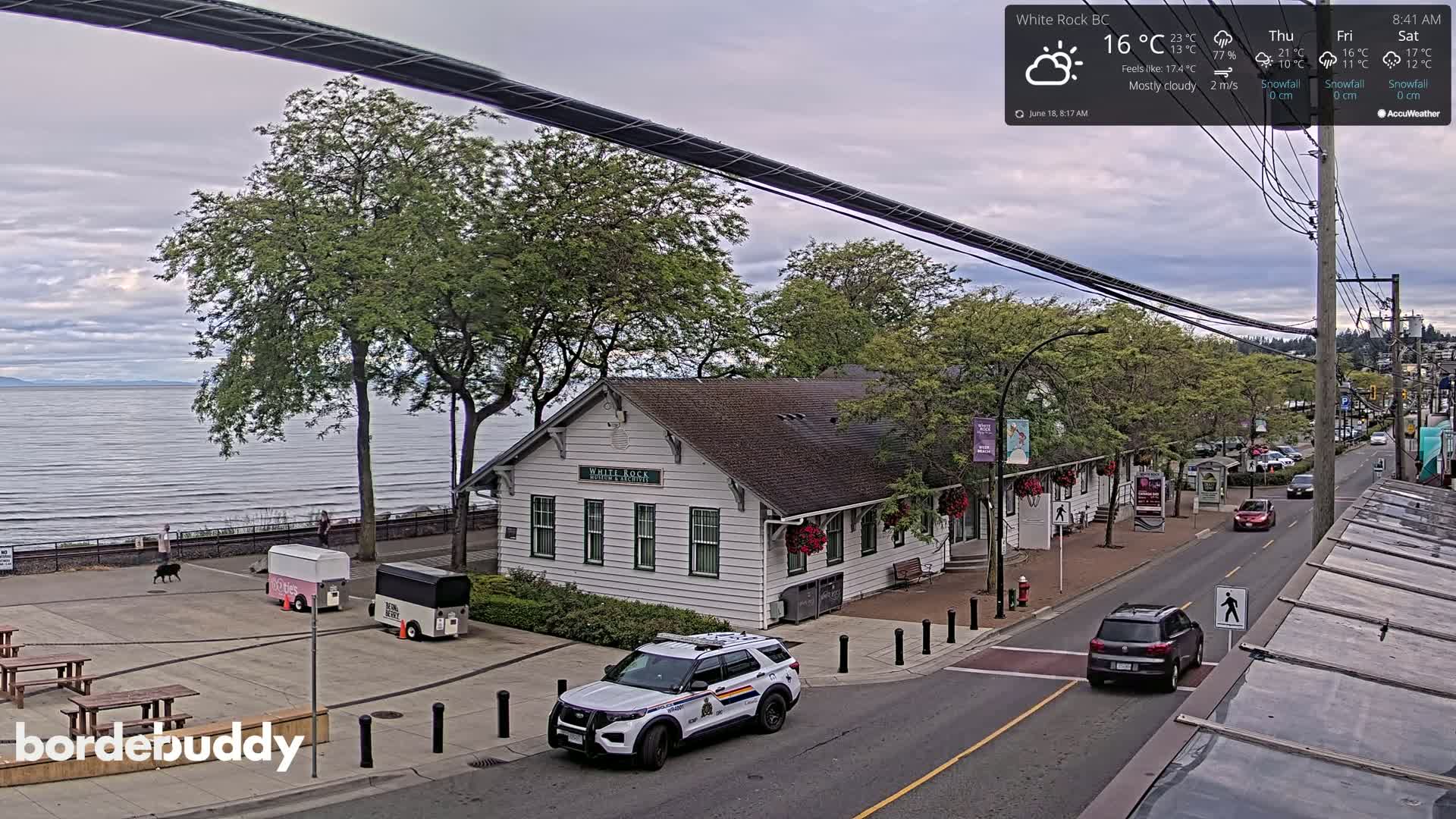 A mostly cloudy day overlooks a waterfront town with a white building, parked food carts, and a police vehicle on a street with cars.