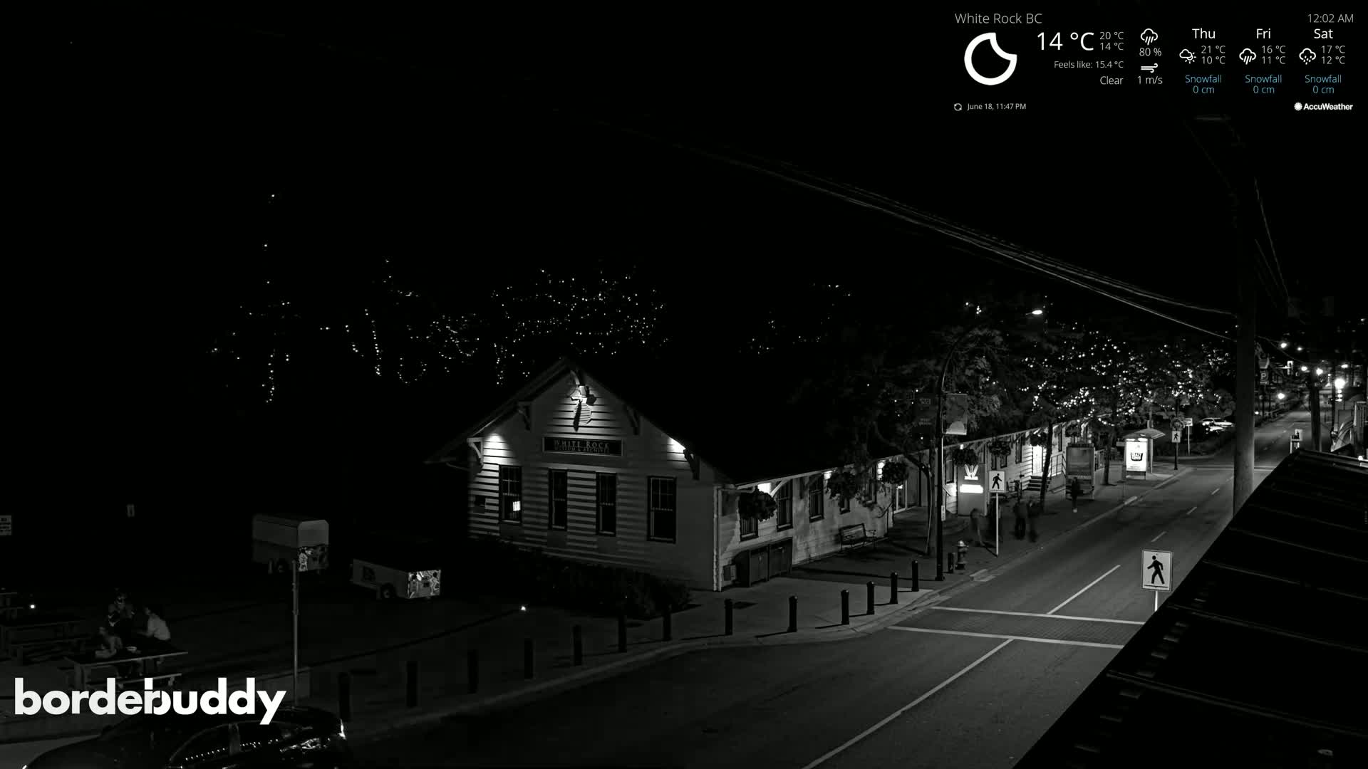 A nighttime, clear view of a street scene with a building illuminated, string lights in trees, and a few pedestrians.