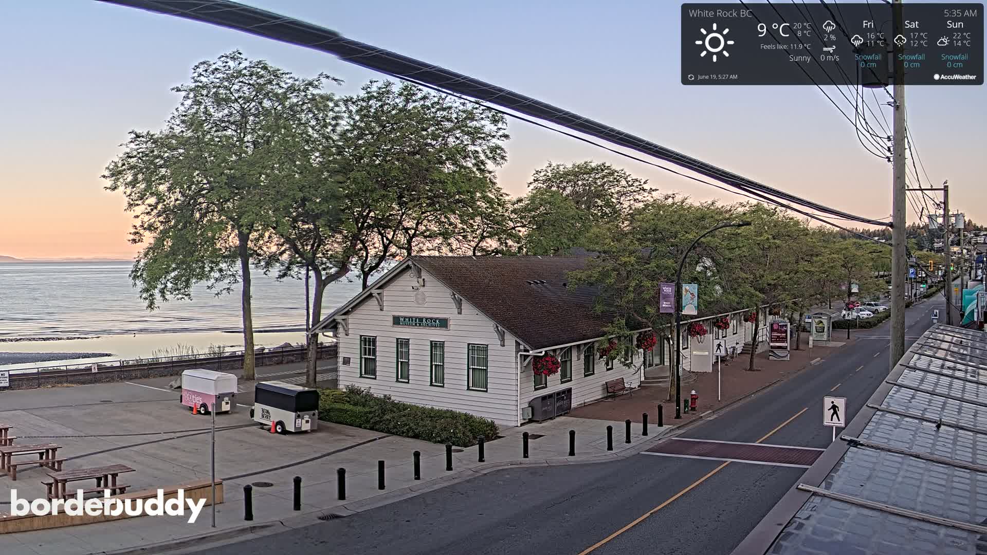 A white building sits near a beach at sunrise under sunny skies, with a street and parked vehicles visible.