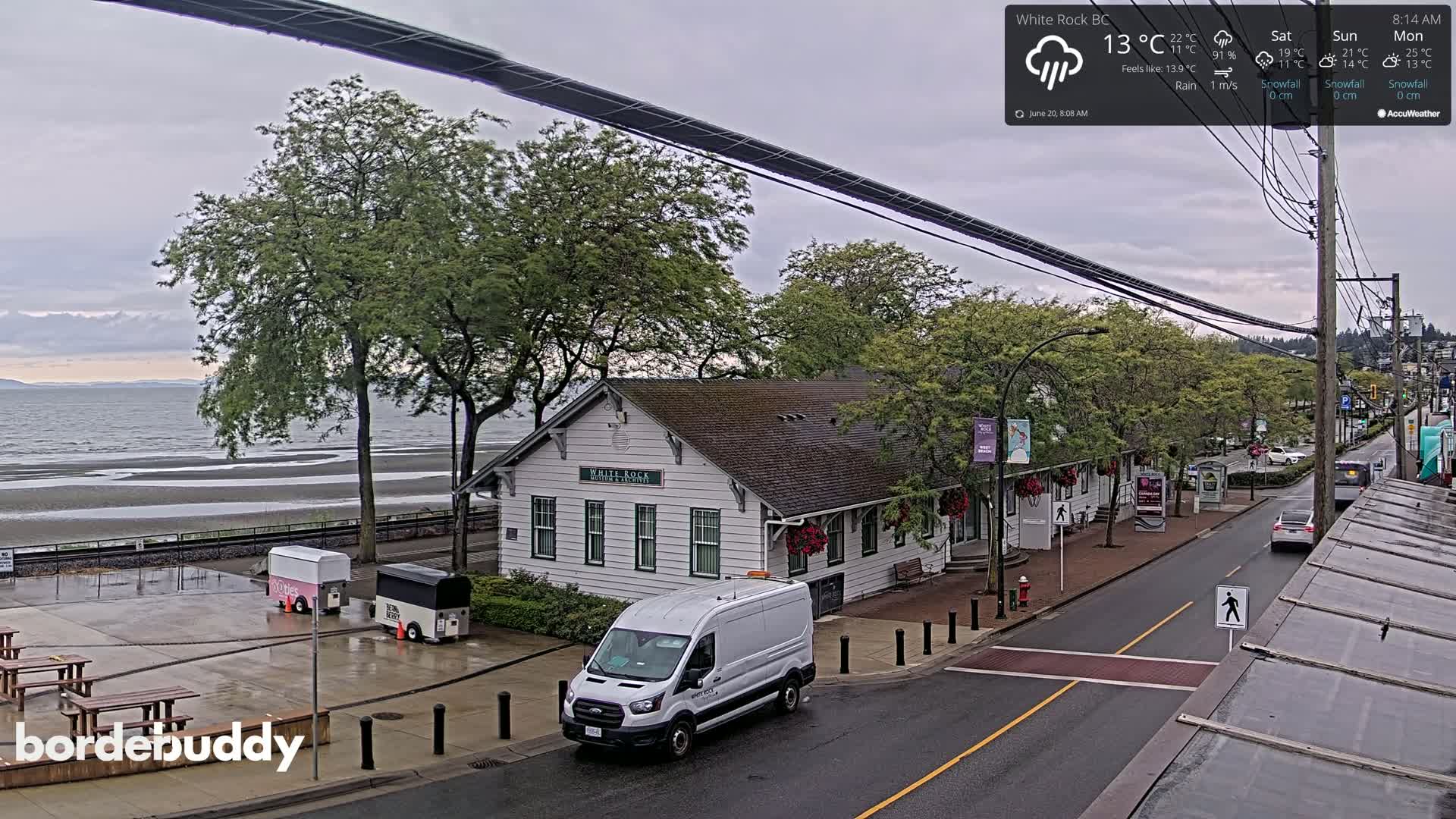 A seaside town scene on a rainy day shows a white building near the shore, a street with vehicles, and a calm ocean in the background.