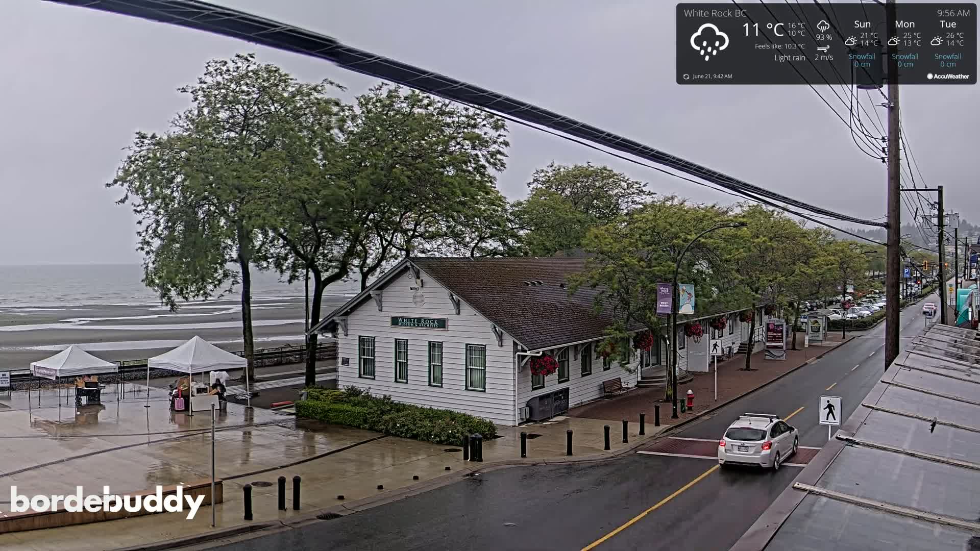 A light rain falls on a seaside town, showing a white building near a beach with some tents and a car driving down a wet street.