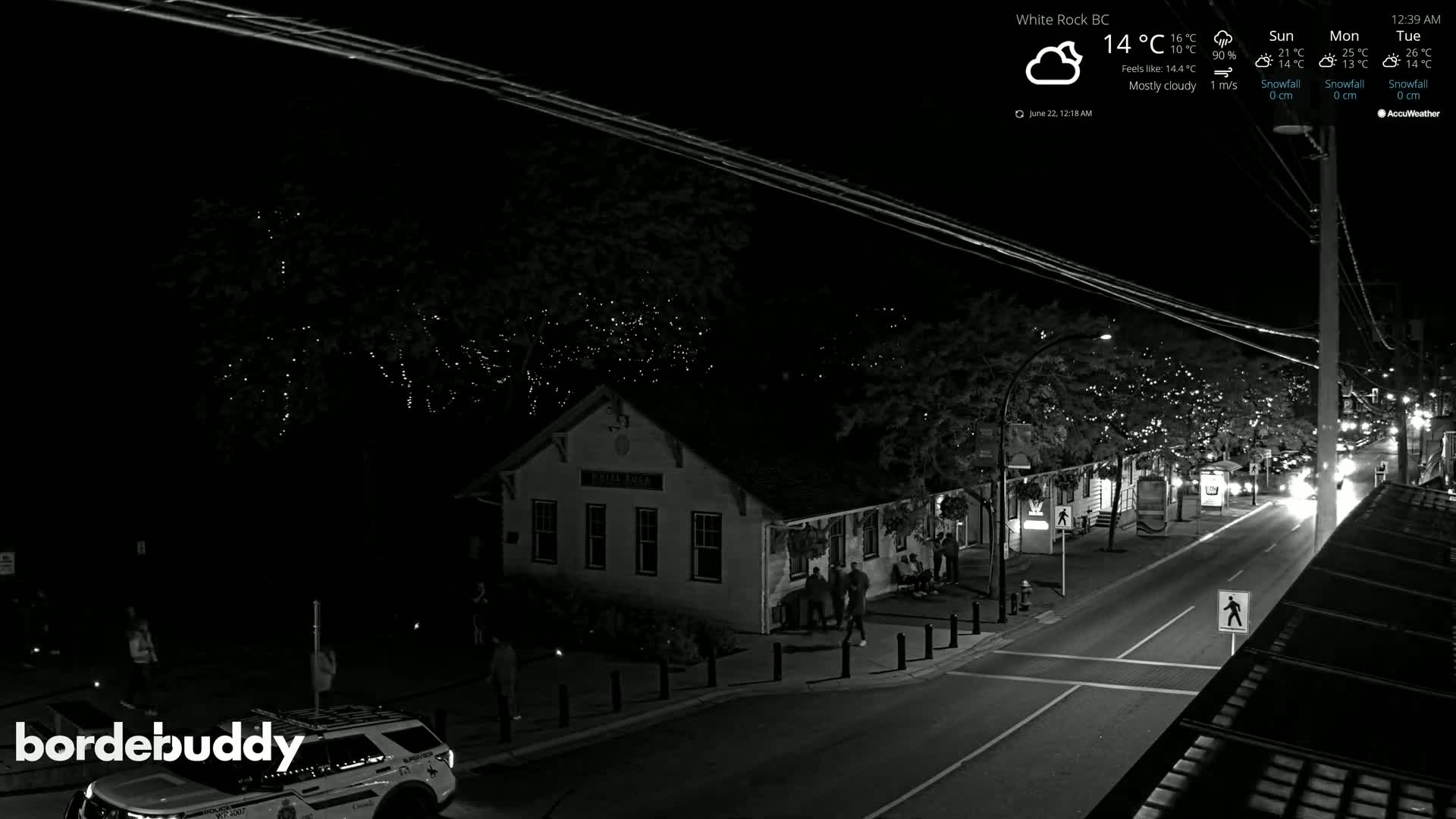 A mostly cloudy night scene shows a street with a building and vehicles, illuminated by streetlights and string lights in trees.
