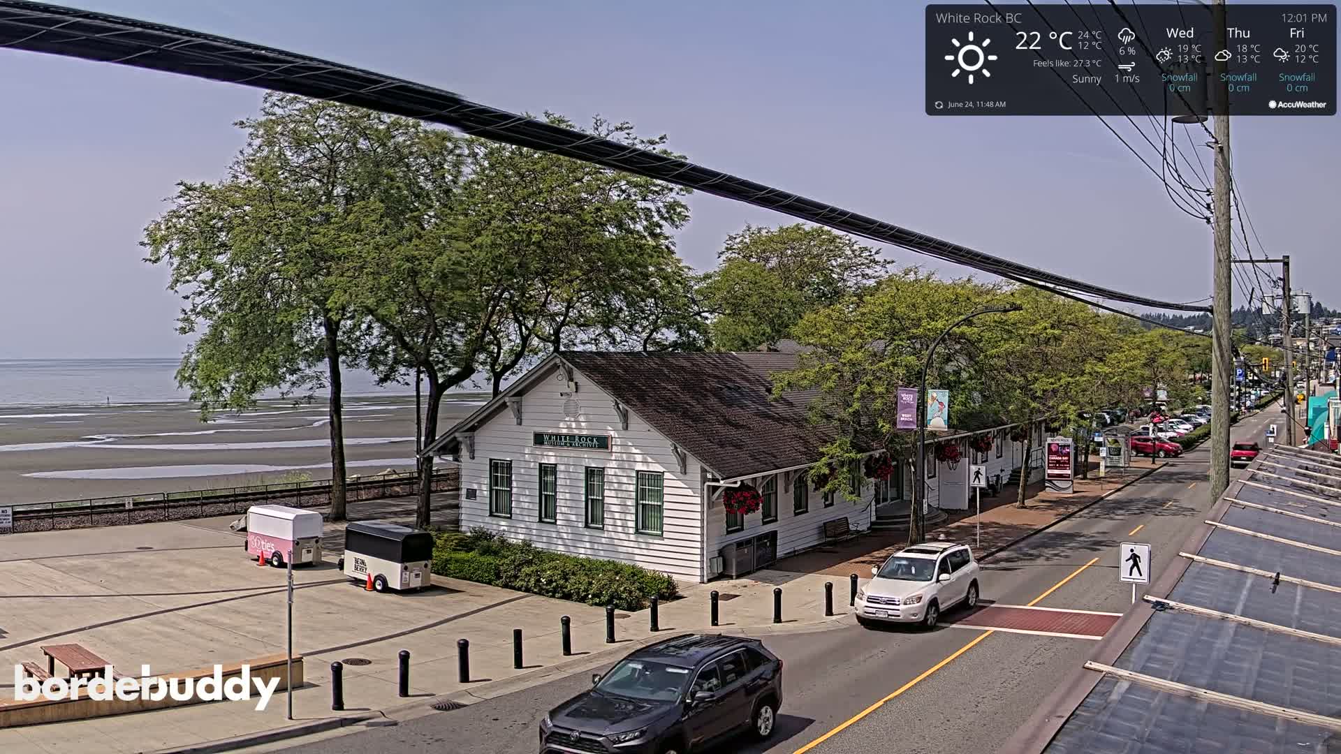 A sunny seaside town features a white building near a beach at low tide, with cars parked along a street and some trees.