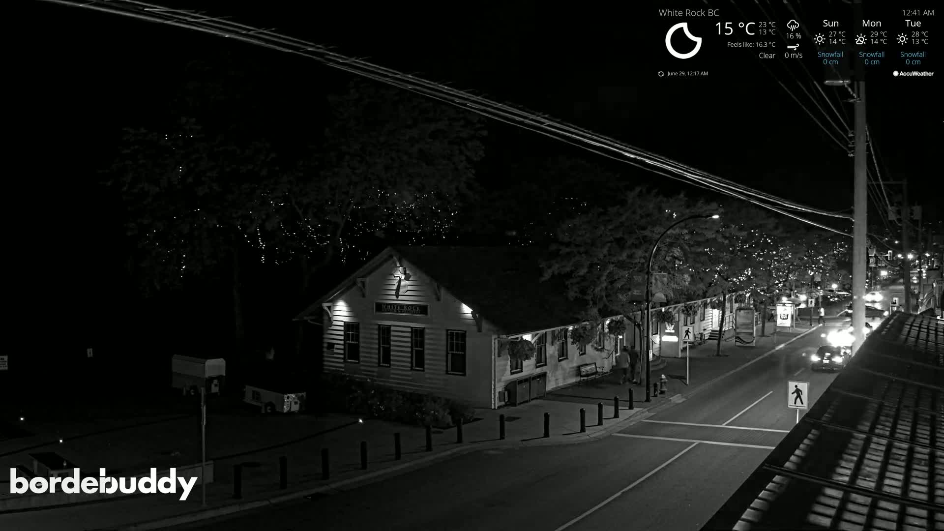 A nighttime street scene shows a building lit up with lights on trees, a road with a car, and a clear sky.