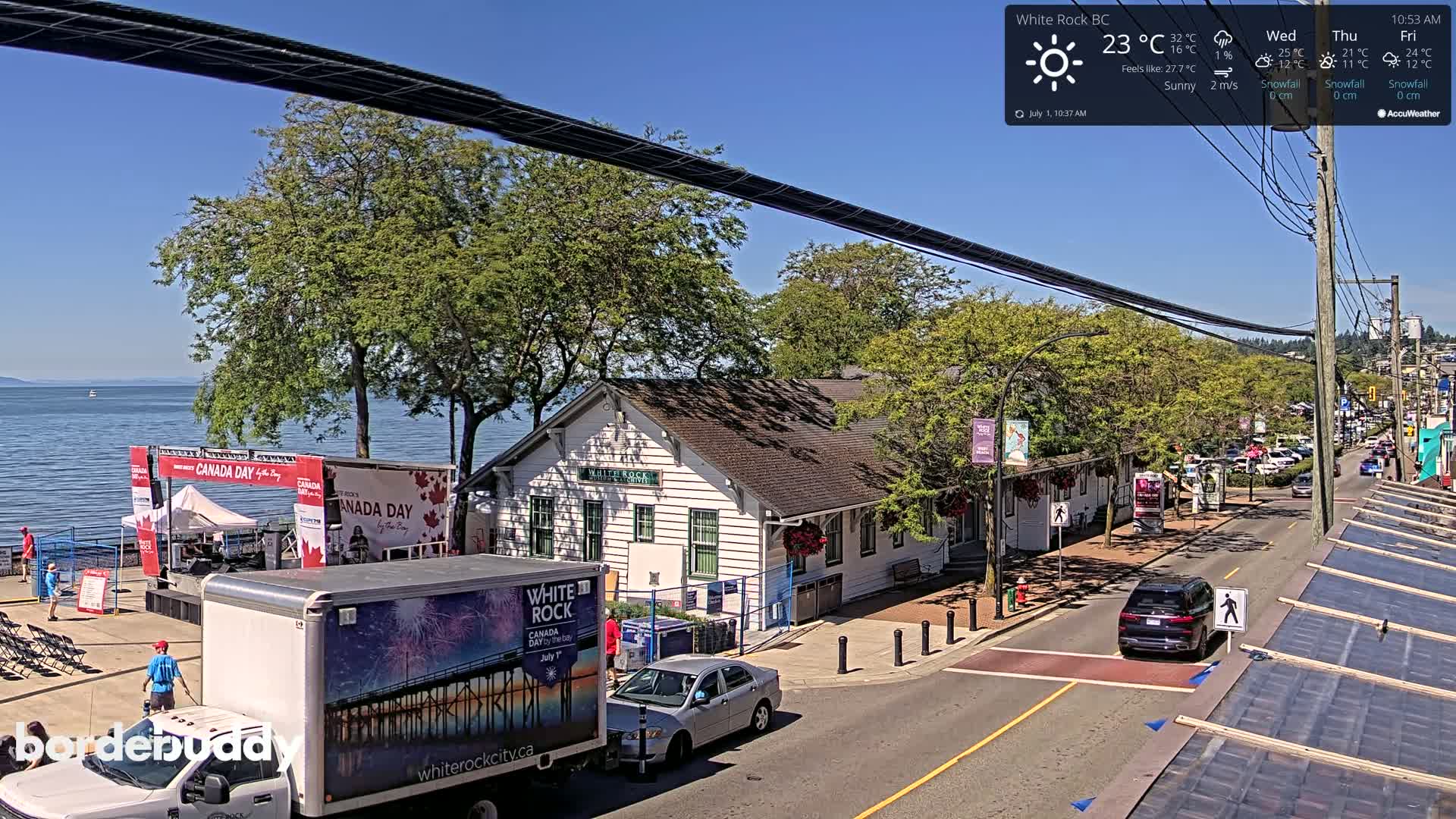 A sunny waterfront scene shows a white building, Canada Day celebrations, parked cars, and a partly cloudy sky.
