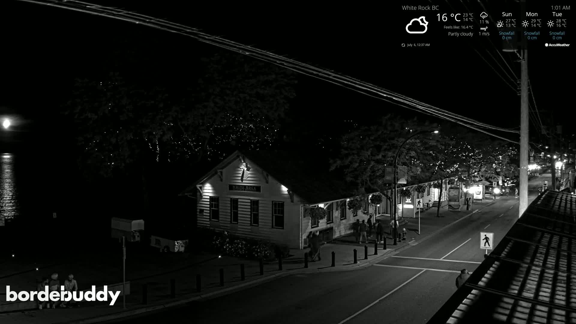 A nighttime, partly cloudy scene shows a street with a few pedestrians near a building by the water, under string lights illuminating trees.