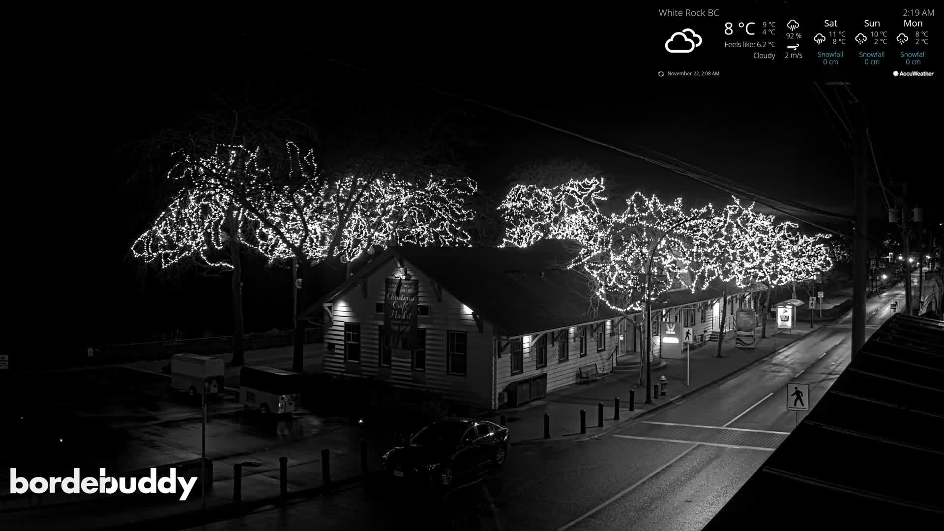 Under cloudy 8°C weather, the image displays a quiet nighttime street lined with buildings and numerous bare trees brightly adorned with white festive lights.